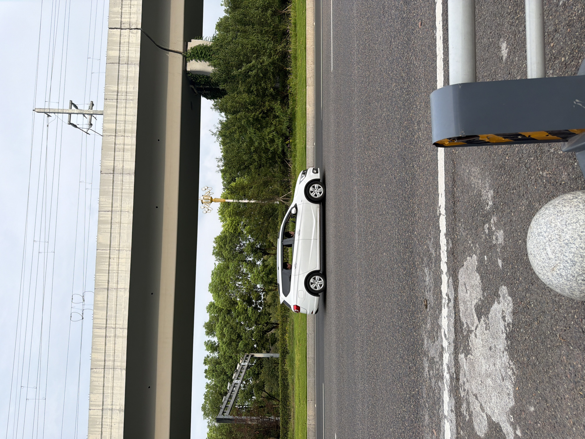 A white station wagon is driving on a paved road under a concrete overpass. The overpass has utility poles and wires above it. There are trees and greenery on the side of the road, with a lamppost visible in the background. The foreground shows the edge of the road with a white line and some decorative concrete spheres and a metal barrier. The sky is overcast. The image appears to be taken from the side of the road, possibly from a sidewalk or median. The vehicle has two occupants visible in the rear seats. The location is identified as Jiangjin, China.