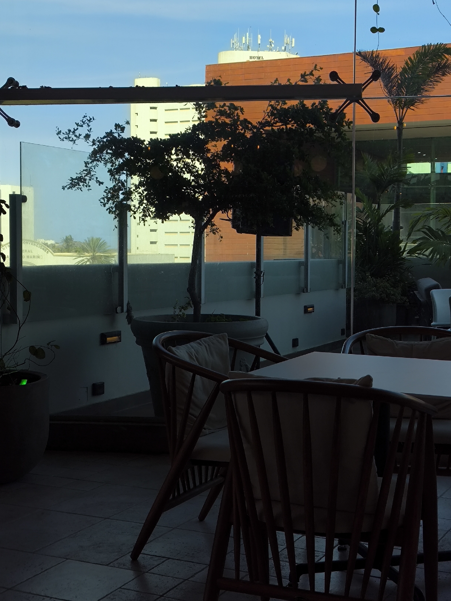 This indoor scene depicts a restaurant or lounge setting, likely a terrace or patio area, in Porlamar, Venezuela. The focus is on the interior elements: wooden chairs and a white table, partially visible, suggesting a dining arrangement. The lighting is dim and subdued, highlighting the texture of the tiled floor and the contours of the furniture.
Through the glass enclosure, one can see a glimpse of the urban landscape. There's a building with a sign that reads "HOTEL", identifiable by the antennas on its roof. Other buildings are also visible, including one with a partially legible sign "CENTRO GENERAL MARINO". A palm tree is seen in the background as well. The overall environment suggests a setting at dusk or in a low-light condition, with the outside cityscape providing a contextual backdrop.