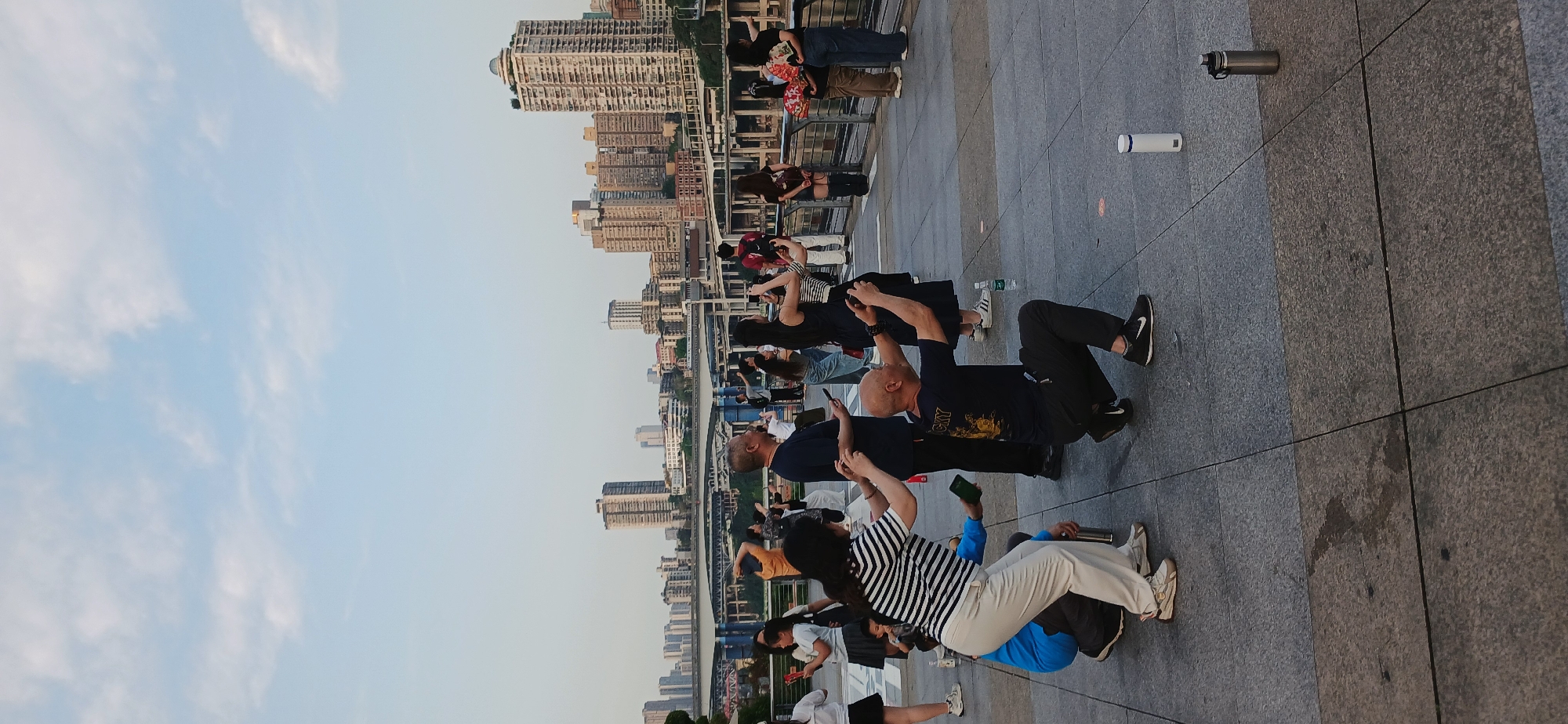 A low-angle, vertically oriented photograph depicts a group of people gathered on a paved surface, with a cityscape visible in the background. The sky is visible at the top of the frame.
The people are of varying ages. They are mostly looking upwards and taking photographs. The people are looking at skyscrapers that are along the background, and along the banks of Jialing River. Water bottles are scattered in the scene.
The location is in the Yuzhong District of Chongqing, China.