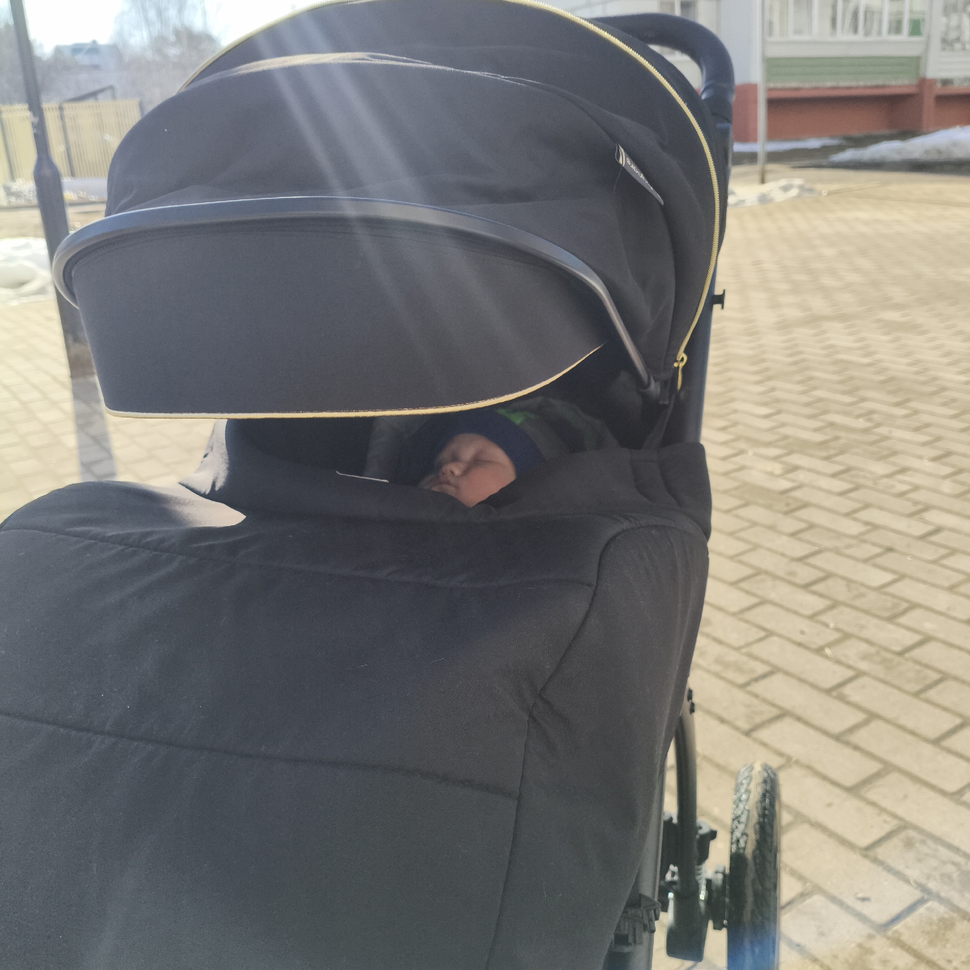 A baby sleeps in a black stroller on a brick sidewalk, likely within the city of Chaikovsky, Russia. A few patches of snow remain on the grass nearby, and a building is visible in the background.