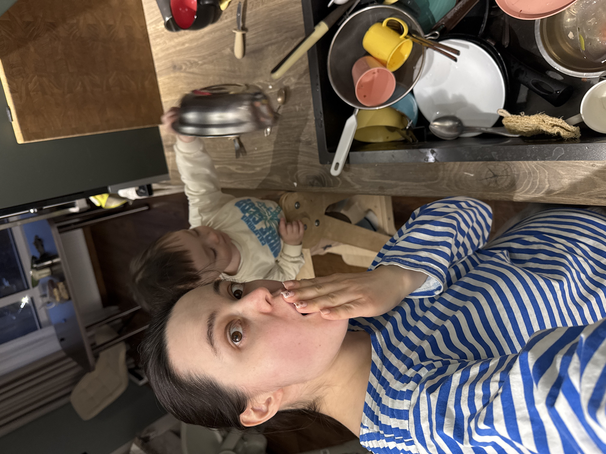 A selfie photograph shows a woman with dark hair, wearing a blue and white striped shirt, holding a hand near her mouth. A young child in a long-sleeved shirt is standing near a kitchen countertop next to the woman. The countertop features a sink full of dirty dishes, including pots, pans, cups, and cutlery. The setting appears to be indoors, possibly in an apartment. No information can be inferred about Izhevsk, Russia, from this image.
