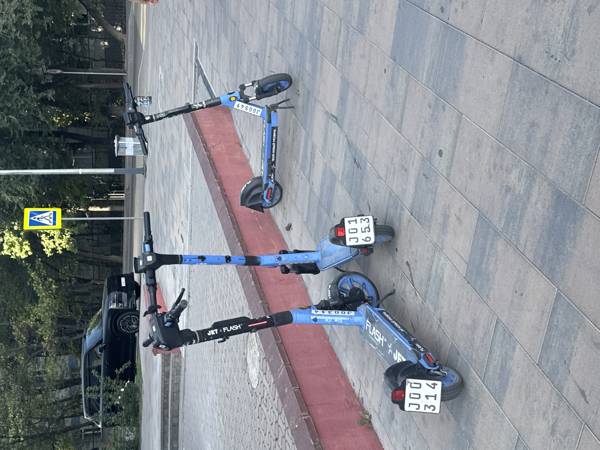 Two blue electric scooters are parked on a paved sidewalk. The scooter on the left has the license plate "JOO 314" and the text "JET X FLASH" and "ТВОЙ НОВЫЙ УРОВЕНЬ" on its deck. The scooter on the right has the license plate "JO1 653" and the number "JO0549" on its handlebar. In the background, a black SUV is parked, and a pedestrian crossing sign is visible. The scene appears to be in Almaty, Kazakhstan, based on the design of the license plates and the overall urban setting.