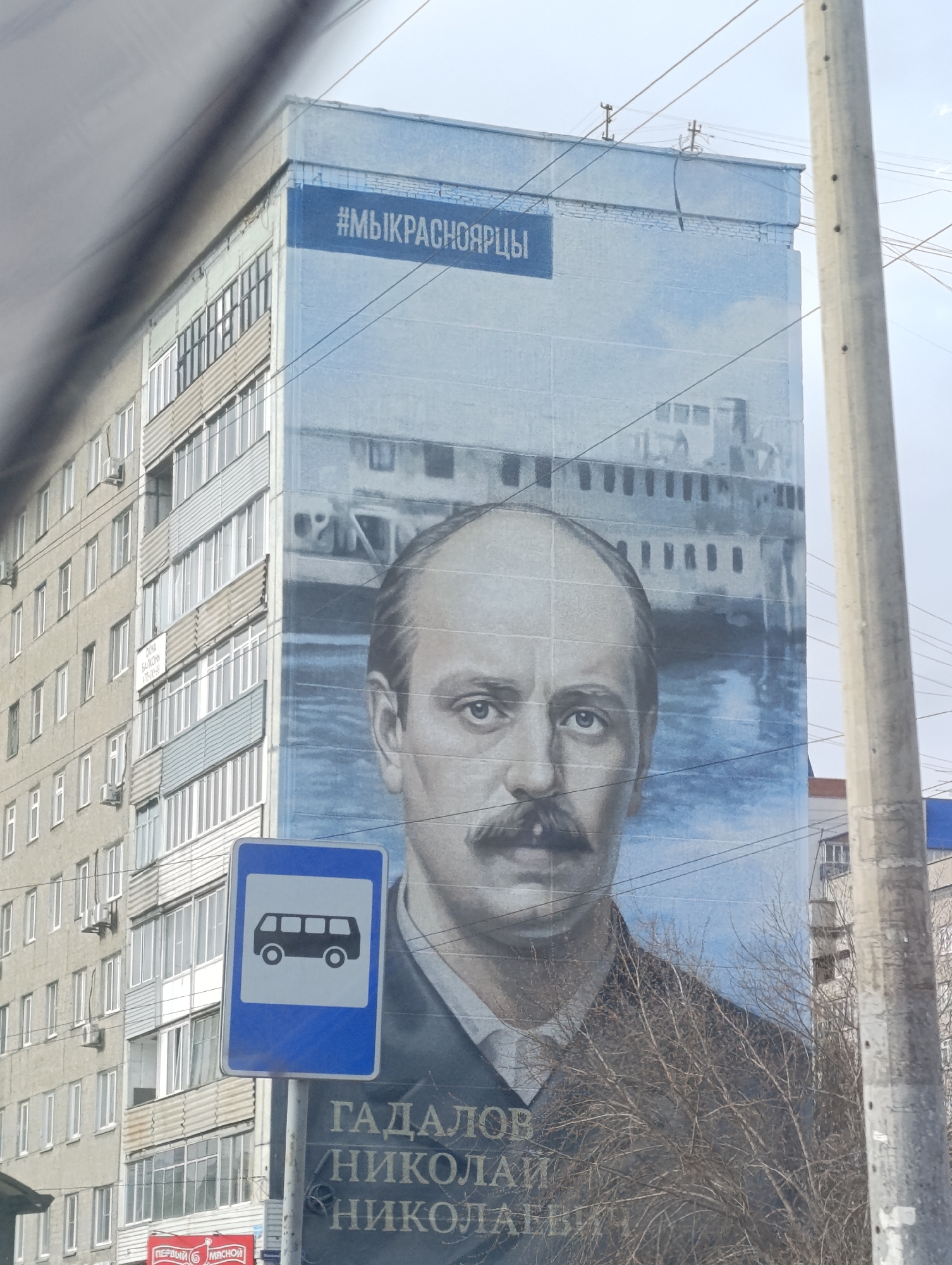 The image captures an urban street scene in Krasnoyarsk, Russia, dominated by a large mural painted on the side of a tall building. The mural features a detailed portrait of a man with a mustache, looking directly forward, dressed in a dark jacket and white shirt. Behind him, a ship is depicted on water, suggesting a historical or maritime context. At the top of the mural, a blue band displays the white text "#МЫКРАСНОЯРЦЫ" (We are Krasnoyarzi/Krasnoyarsk residents). Below the man's portrait, his name is written in white capital letters: "ГАДАЛОВ НИКОЛАЙ НИКОЛАЕВИЧ" (Gadalov Nikolai Nikolaevich).
Adjacent to the mural, another multi-story residential building with numerous windows, balconies, and air conditioning units is visible. In the foreground, a prominent blue rectangular bus stop sign with a white bus icon stands on a pole. Bare tree branches partially obstruct the view of the mural and the bus stop sign, indicating a season like late autumn or winter. Overhead, multiple power lines are visible against an overcast sky, suggesting it is daytime. A small, blurry sign with unreadable text is attached to the side of the adjacent building. Below the bus stop sign, a partial red shop sign reads "ПЕРВЫЙ МЯСНОЙ" (First Meat). The scene presents a blend of public art and everyday city life.
