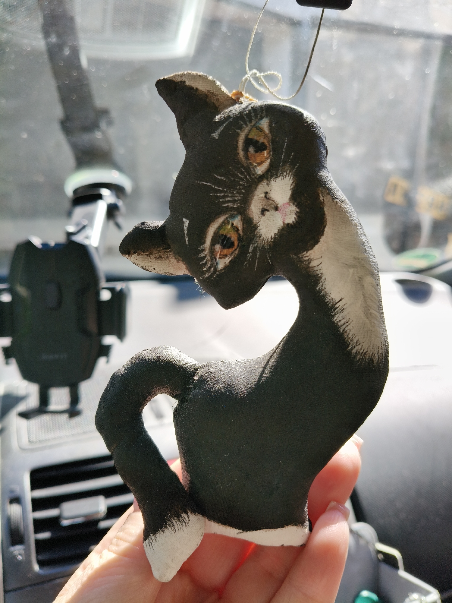 A black and white cat-shaped ornament is centrally depicted, held by a human hand. The ornament features a dark, textured body with white accents on the ear tips, nose bridge, mouth area, chest, and front paws. It has prominent, multi-colored eyes displaying shades of yellow, green, and brown, with black pupils. A thin, light-colored string is tied to the top of the ornament's head, indicating it is designed to be hung. The ornament's tail forms a distinct heart shape. A light-skinned human hand, with visible fingernails, supports the lower portion of the ornament. The background reveals the interior of a vehicle, likely a car, under bright, possibly sunny, conditions. Visible elements in the blurred background include a black car phone mount positioned to the left, a car air vent with horizontal slats on the lower left dashboard, and indistinct reflections and light flares on what appears to be a windshield.