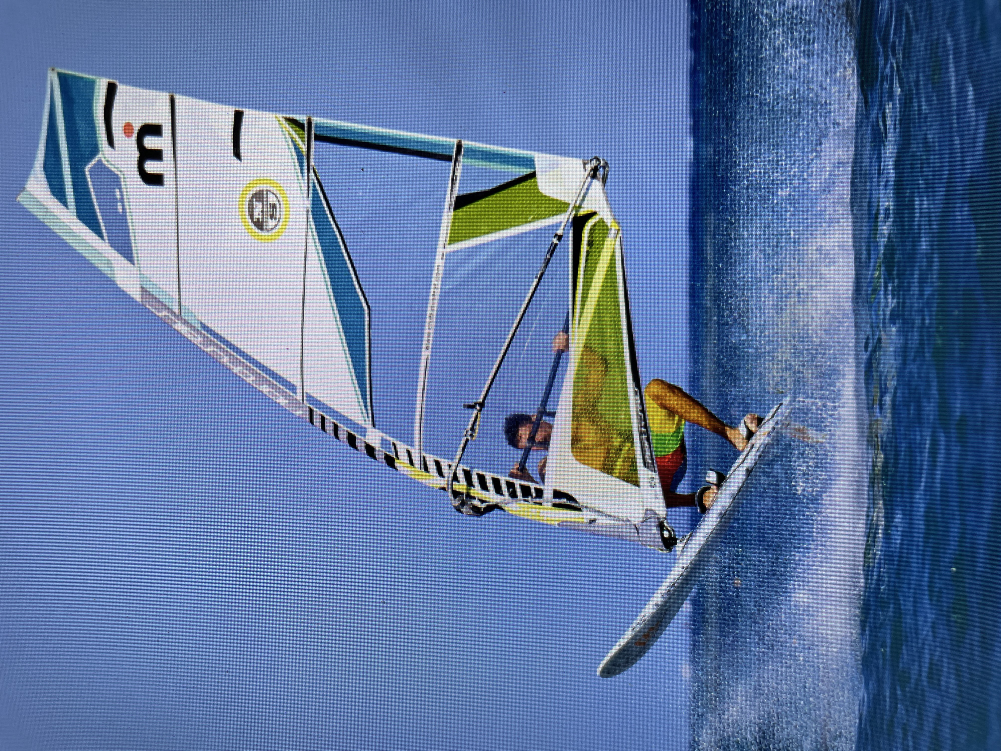 A windsurfer is in action. The sail is white with blue and green accents, displaying a large "3.1" and a circular logo. The windsurfer, wearing a yellow shirt and patterned shorts, holds onto the boom. The board is white. Water spray indicates movement. The background consists of a blue sky and ocean. The location is likely coastal, possibly in Bacong, Philippines.