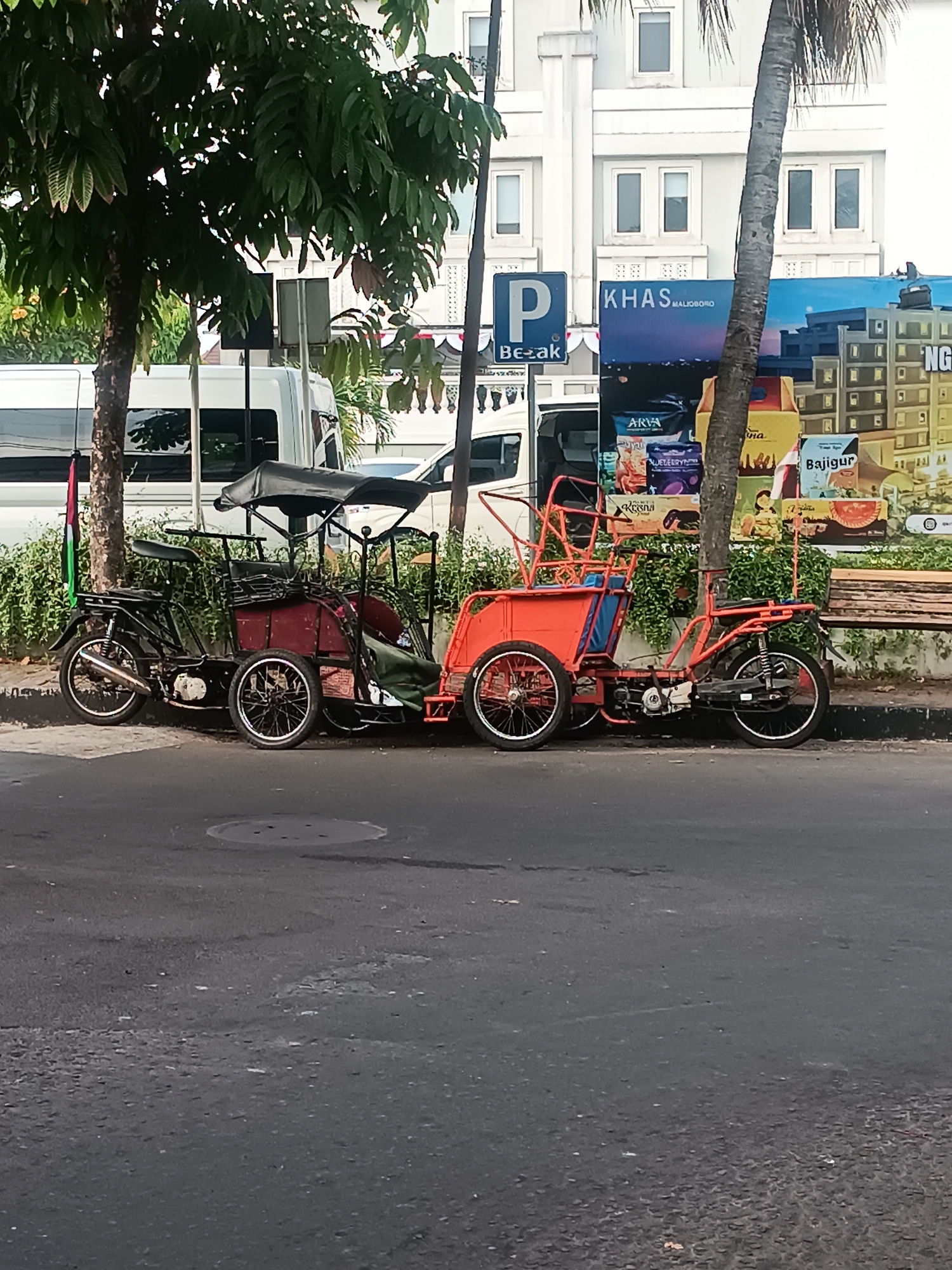 The image depicts a street scene in Yogyakarta, Indonesia, likely near Malioboro street, evidenced by a sign reading "KHAS MALIOBORO". In the foreground, two colorful rickshaws, or "becak", are parked on the side of an asphalt road. One is black with a red passenger compartment and a black canopy, while the other is entirely bright orange. Both have three wheels and appear to be motorized. Behind the rickshaws, a white van is visible, and further back, a modern building with multiple windows stands. Lush green foliage from trees and bushes lines the sidewalk, adding a natural element to the urban landscape. The overall atmosphere suggests a typical day in a bustling Indonesian city.