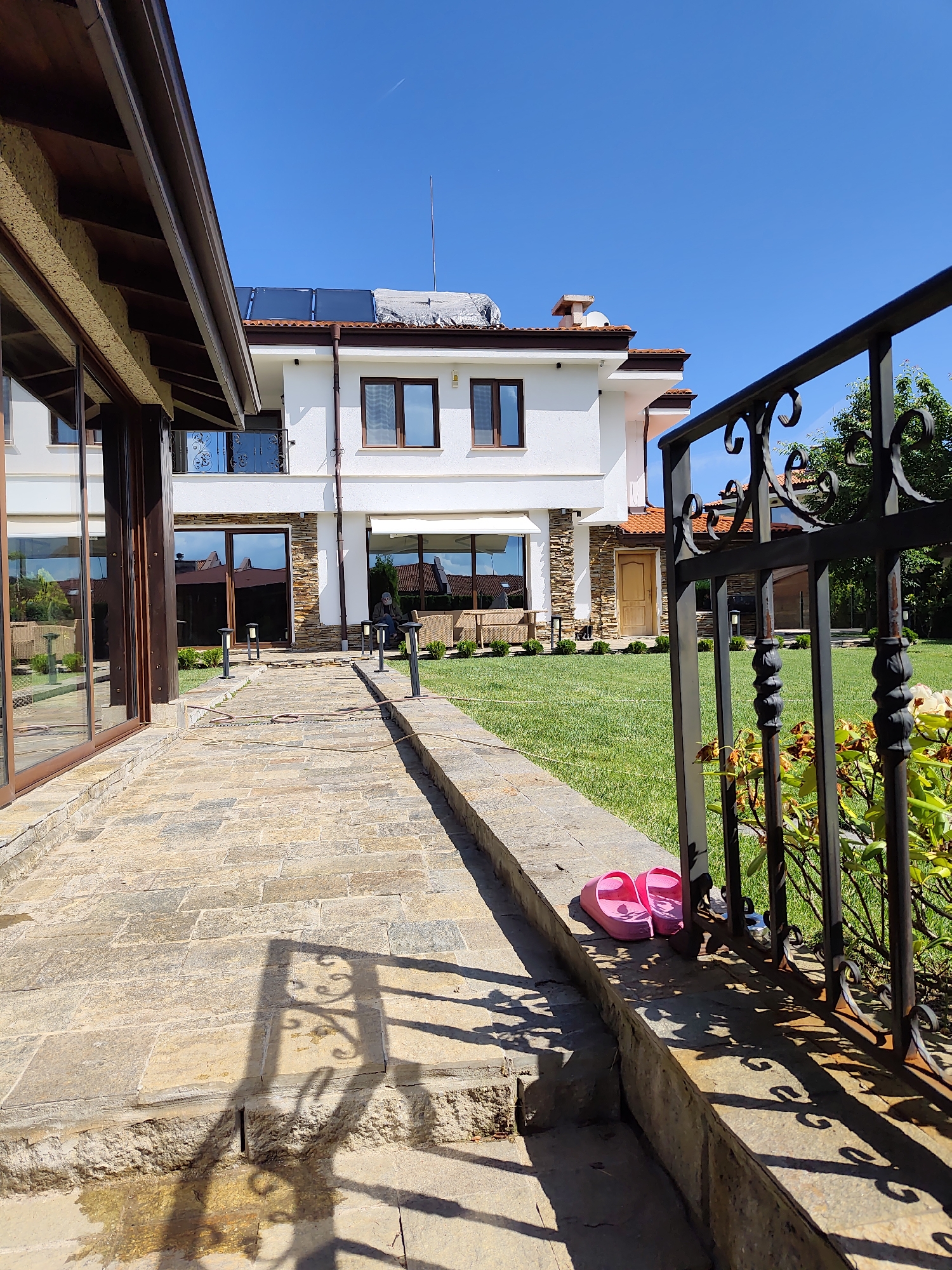 The image depicts a sunny day in Kladnitsa, Bulgaria, with a focus on the exterior of a modern residential property. A stone pathway leads towards a two-story white house with dark-framed windows and a tiled roof. Solar panels are visible on the roof of the house. To the left of the pathway, a portion of another building with a brown roof and large glass windows is visible. The yard is covered in green grass, and a black wrought-iron fence runs along the right side of the image. A pair of pink flip-flops rests on a stone ledge in front of the fence. The environment is well-maintained and suggests a peaceful, residential setting.