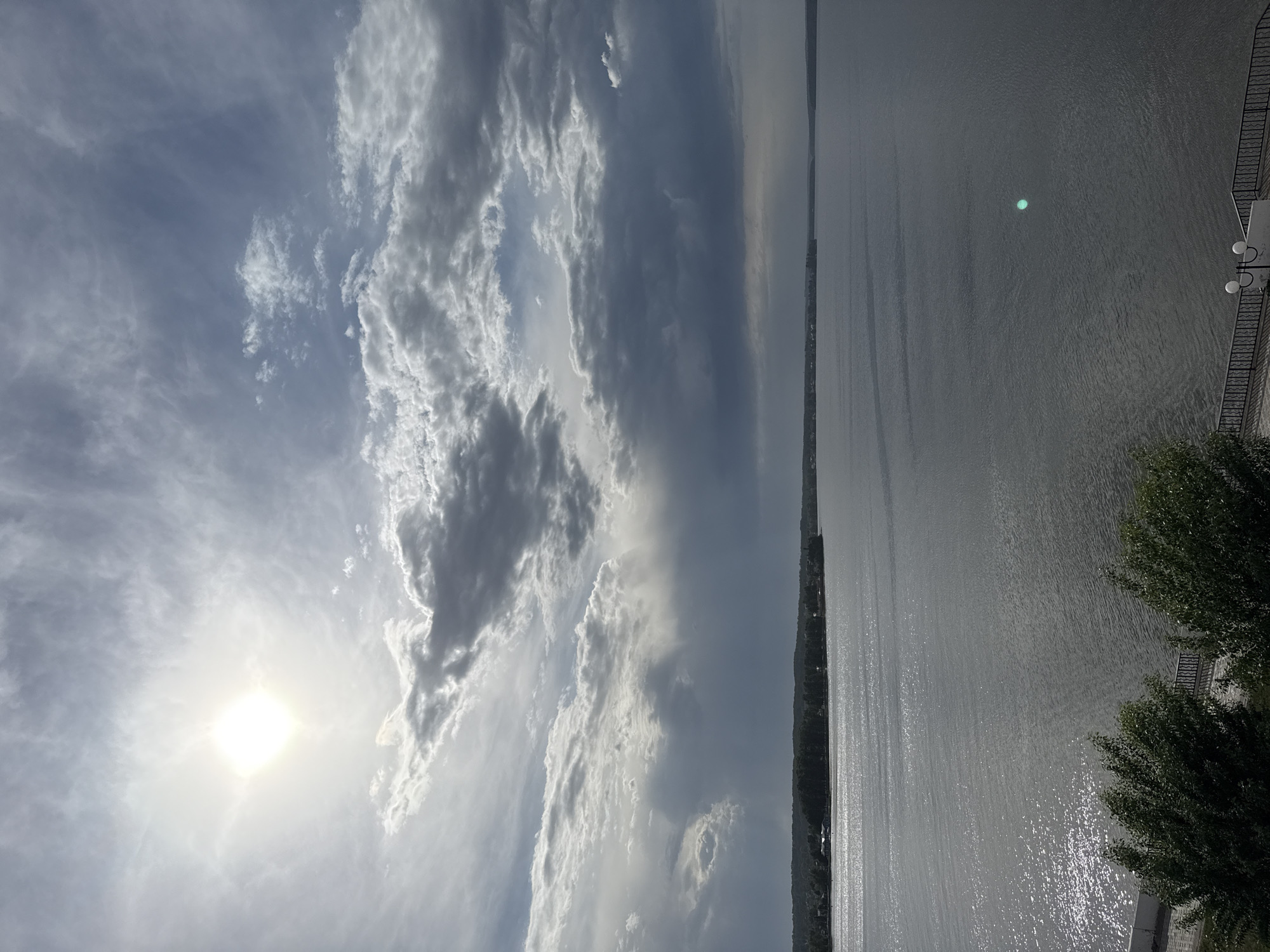 The image captures a landscape featuring a body of water under a sky filled with diverse cloud formations. The sun is visible and prominent, partially obscured by the clouds. A stretch of land, possibly a shoreline or bank, is situated along the water's edge, with some vegetation visible along the border. On the right side, there's a suggestion of a built environment, perhaps a street or a pathway, with structures that could be streetlights or architectural features. The water surface reflects sunlight, creating a shimmering effect.
This scene is likely located in Votkinsk, Russia, on a river or lake with a developed embankment.
