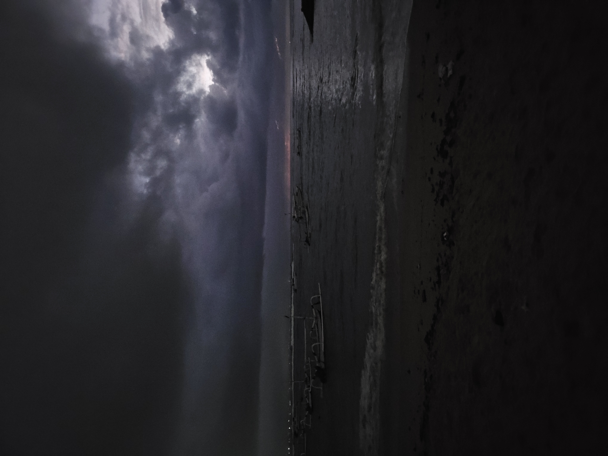 The image depicts a dark, overcast scene at what appears to be Sanur Beach in Denpasar, Indonesia. Storm clouds dominate the sky, with a small patch of light peeking through. The sea is calm, reflecting the dim light. Several traditional outrigger boats, known as "jukung," are visible on the beach and in the water. The beach itself is dark, with visible sand. The image is taken at night or during twilight, adding to the somber atmosphere. No people are visible in the scene.