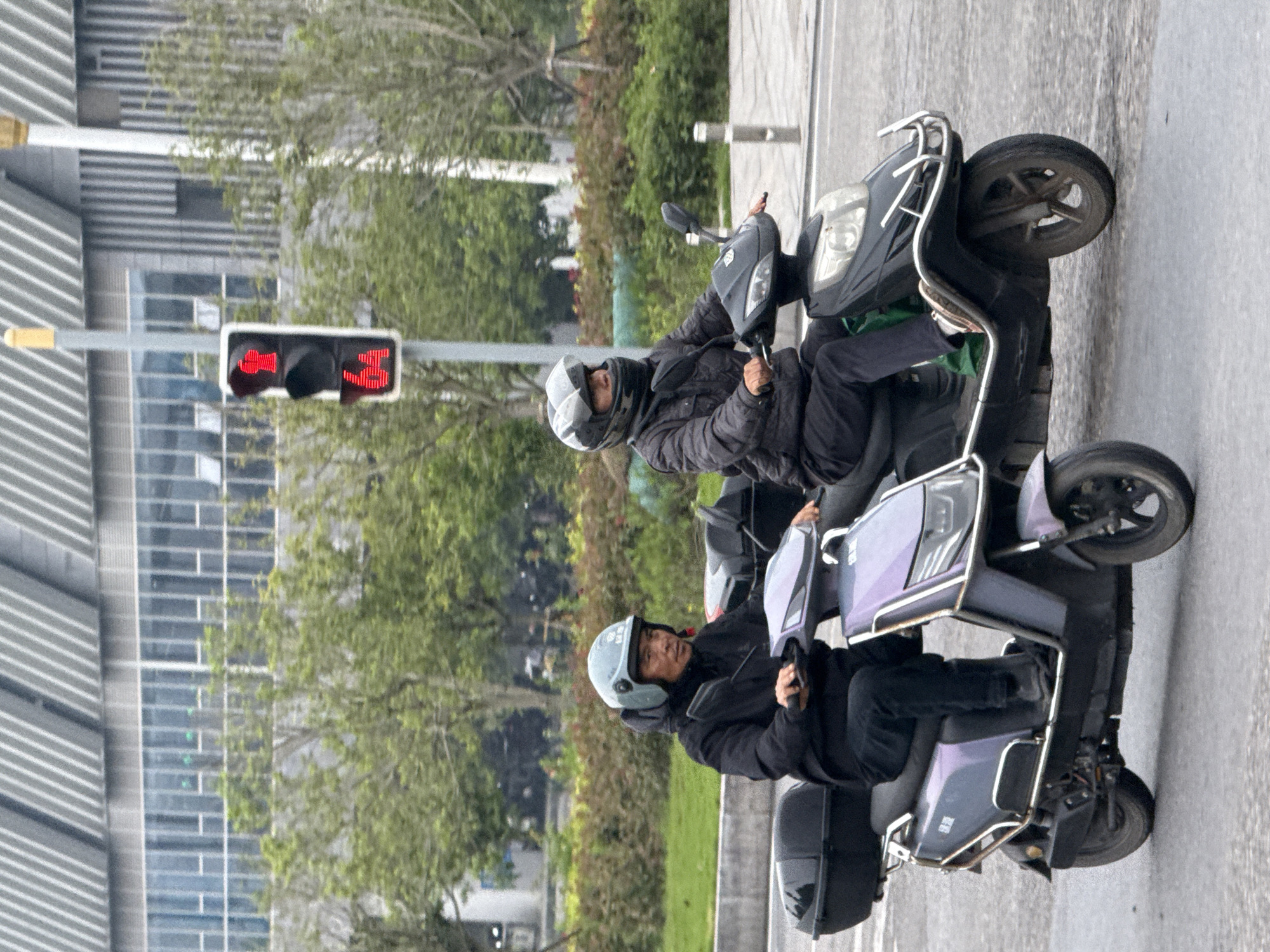 Two men in helmets are riding scooters at a red light in Jiangjin, China. The light shows a countdown timer with 3 seconds remaining. The buildings are modern, and the area is green with grass and trees.