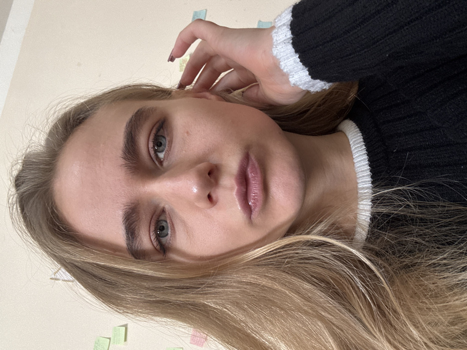 The image captures a frontal, chest-up portrait of an adult female subject indoors, located in the city of Unknown, Unknown. She possesses long, wavy blonde hair, blue eyes, and light complexion. The subject wears a black ribbed sweater featuring a white crewneck collar and visible white cuffs on her right arm. Her lips are full, glossy, and slightly pursed. Her right hand is raised, fingers lightly contacting the right side of her head near the ear; her fingernails are painted dark red. The background comprises a plain, light-colored wall with several small, square sticky notes in green, pink, and light blue hues affixed to it. A faint drawing is discernible on the upper left portion of the wall. The subject maintains direct eye contact with the viewer.