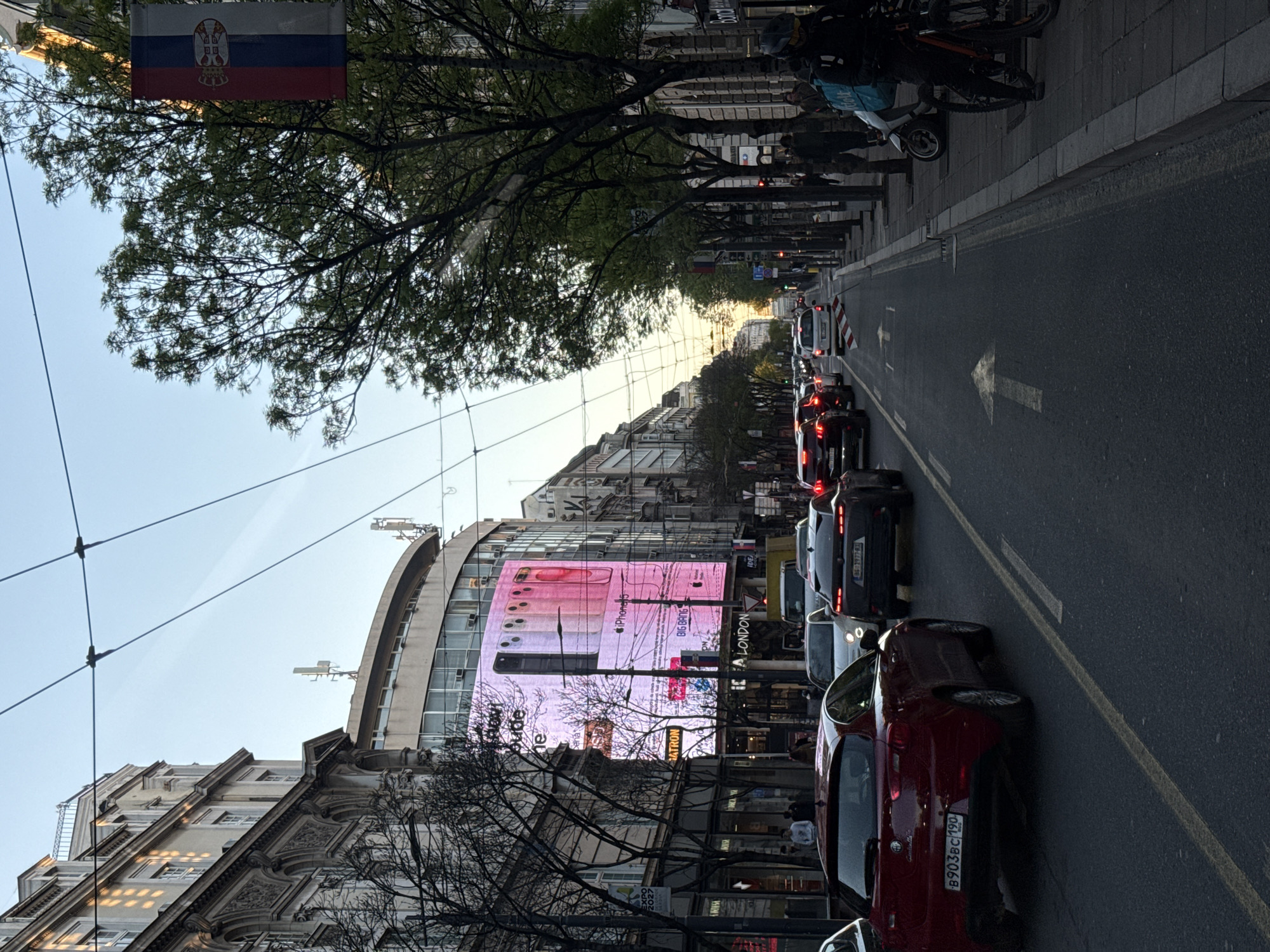The image shows a street in Belgrade, Serbia, filled with stationary cars. A large billboard displays advertisements, including a prominent iPhone ad. The flag of Serbia is seen above the trees. Pedestrians walk on the sidewalks along the sides of the street. An arrow is painted on the road surface. Delivery drivers are seen with scooters. Overhead wires are visible against the sky.