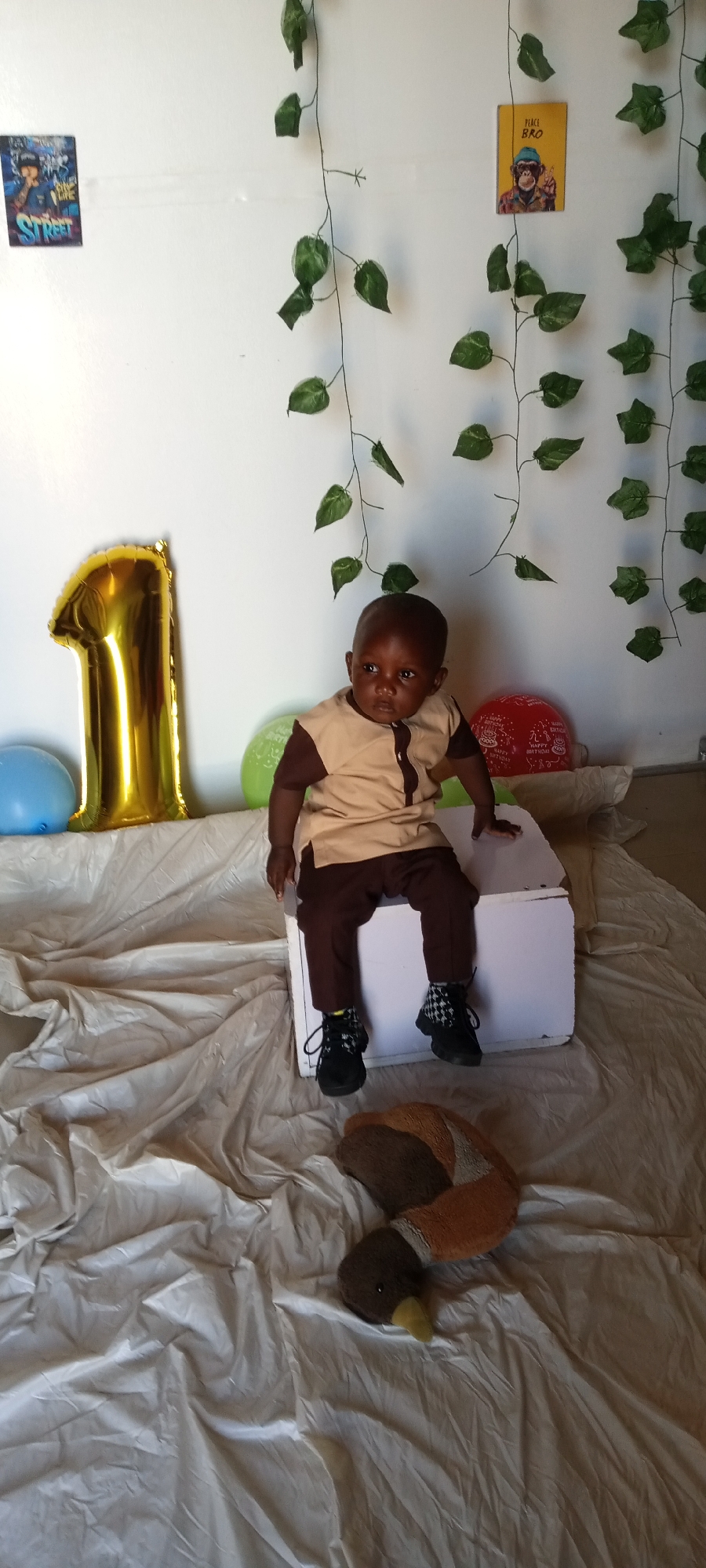 The image captures a child, presumably around one year old, seated on a white box. The child wears a brown and beige outfit paired with black shoes and patterned socks. The backdrop consists of a white wall adorned with faux ivy strands, a golden number "1" balloon, and several colored balloons. Two small posters are affixed to the wall: one depicting an urban scene and the other featuring a monkey with the inscription "Peace Bro". A plush duck toy rests on a wrinkled white cloth laid out on the floor. The scene appears to be a staged setup for a birthday photoshoot. There is no definitive way to establish the exact location of the scene within Jos, Nigeria from visual data.