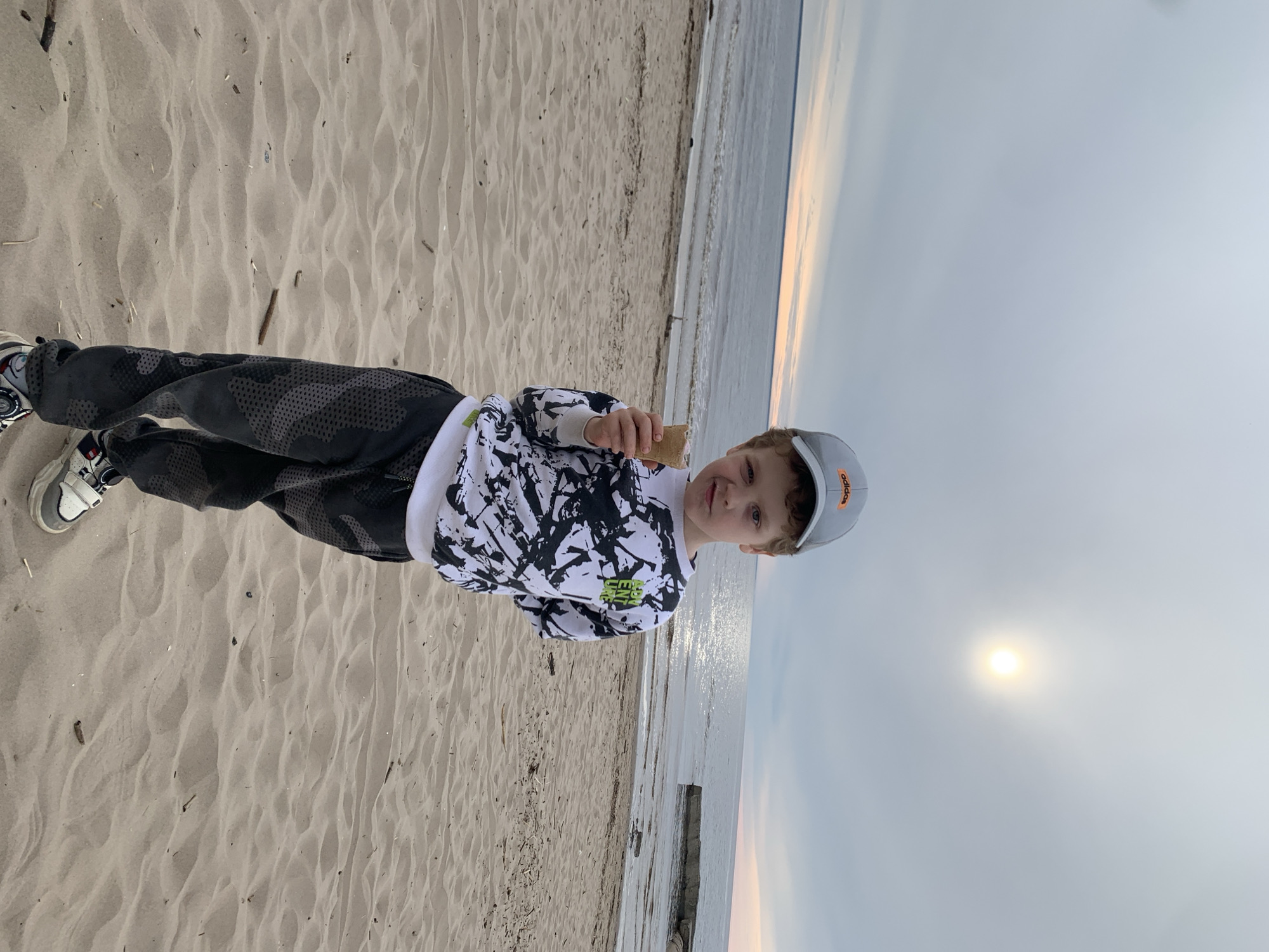 A young boy is standing on a sandy beach with the sea visible in the background. He is wearing a light-colored Adidas cap, a long-sleeved black-and-white patterned shirt, and dark pants with a textured design. The boy is holding a small brown object, possibly a snack, in his hand. The sky is overcast with the sun barely visible through the clouds. The sandy beach appears to stretch out into the distance. There is driftwood visible on the beach. The specific location is difficult to determine from the image but is located somewhere within the city of Severodvinsk, Russia, because that's where the picture was taken.
