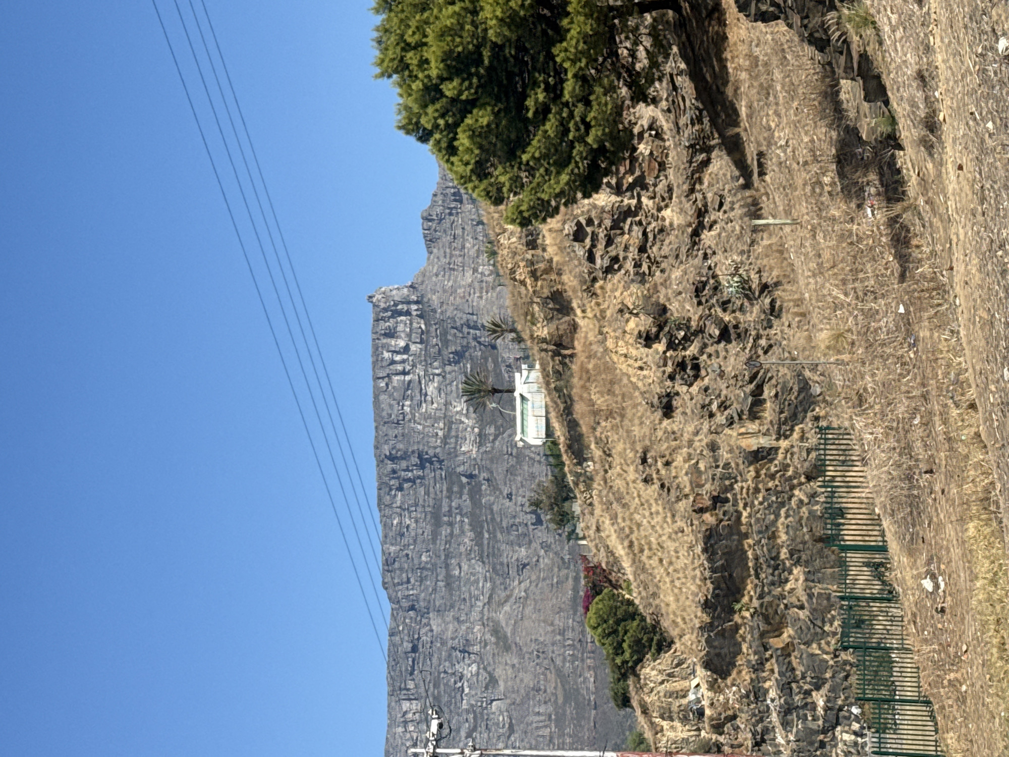 The image depicts Table Mountain in Cape Town, South Africa, under a clear blue sky. In the foreground, there is a rocky hillside covered in dry vegetation and interspersed with some greenery. The mountain itself is a massive, gray, flat-topped formation. There's a building with white walls and a palm tree next to it, situated partway up the mountain's slope. Overhead power lines are visible in the upper left corner of the image. A green metal fence runs along the edge of the hillside. The picture is oriented vertically.