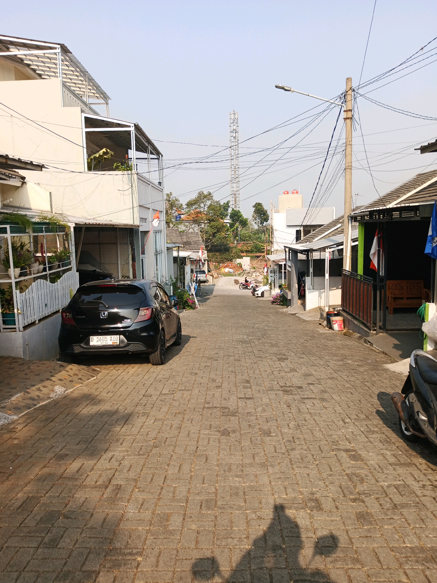 The image depicts a street scene in Bandung City, Indonesia, under a clear sky. The street is paved with interlocking brick pavers and is lined with buildings on both sides. On the left, a two-story building with a white facade and multiple balconies is visible. A black Honda hatchback with license plate D 1605 AID is parked on the left side of the street. On the right, a row of single-story structures with pitched roofs and awnings runs along the street, some with outdoor seating areas and potted plants.
Further down the street, a tall transmission tower stands in the distance. Several utility poles with a complex network of electrical wires crisscross the scene. Two motorcycles are partially visible, one on the right side of the street and another parked further up the street. The overall impression is that of a residential or mixed-use area in an urban environment. Shadows of people and possibly a vehicle are cast on the brick-paved street, indicating the presence of sunlight.