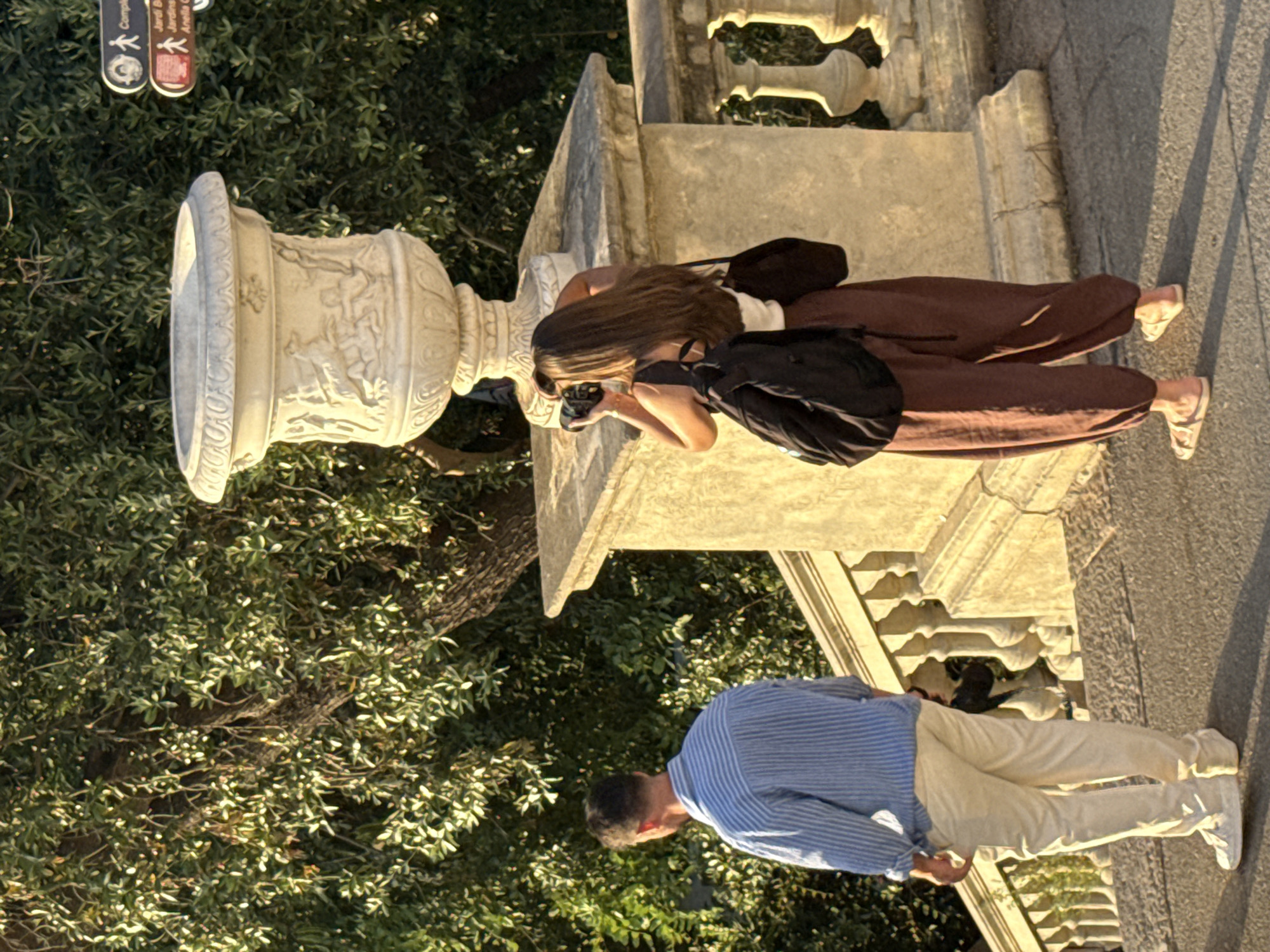 The image depicts two individuals on a stone staircase with ornate balustrades and large stone planters. The setting is illuminated by the warm light of what appears to be late afternoon sun, casting long shadows. The background is filled with lush green foliage from trees.
On the left, a man is viewed from the back, wearing a light blue and white striped shirt and light-colored pants. He is standing on the stairs, facing away from the camera.
On the right, a woman is also seen from the back, holding a camera to her face as if taking a photograph. She has long, dark hair and is wearing a white top with dark, loose-fitting pants and sandals. A black backpack is slung over her shoulders, and a dark bag is hanging from her right arm.
In the background, a large, decorative stone urn sits on a pedestal. To the right of the urn, a signpost with directional arrows and text, seemingly in Catalan, indicates "Compleix", "Jardí Botànic", and "Anella Verda". These signs, particularly the mention of "Jardí Botànic" (Botanical Garden), strongly suggest the location is within Barcelona's Parc del Castell de Montjuïc, specifically near the entrance to the Botanical Garden. The architecture of the balustrades and the urn are consistent with the style found in this historic park.