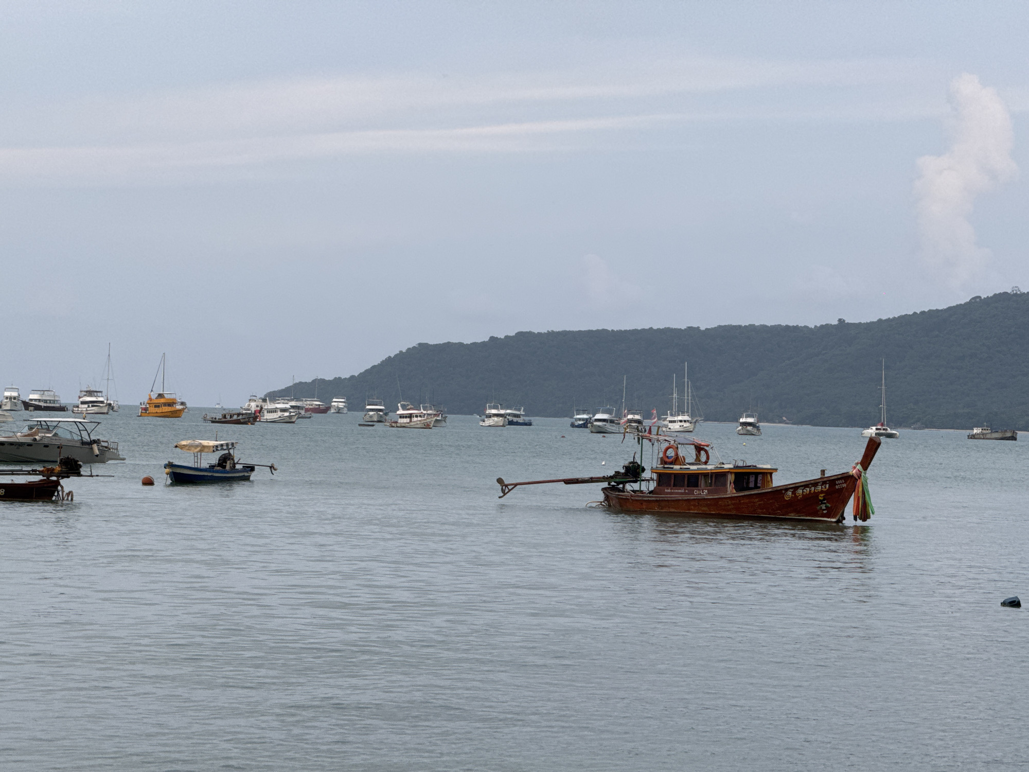 A wide shot captures a tranquil scene in the waters of Chalong, Thailand. The sky is overcast with a soft, diffused light, casting a muted tone over the landscape. In the foreground, the calm, grey water is dotted with various boats. A prominent wooden longtail boat with an extended propeller shaft is positioned in the mid-right of the frame, its hull bearing the inscription "CH-L21" and Thai script. To its left, a smaller blue and white boat with a canopy shelters a lone figure. Further back, numerous other vessels, including sailboats and motorboats, are anchored or moored. The background is dominated by a lush, green, tree-covered hill rising from the water's edge, creating a serene natural backdrop to the harbor. The overall impression is one of a peaceful maritime setting under a cloudy sky.