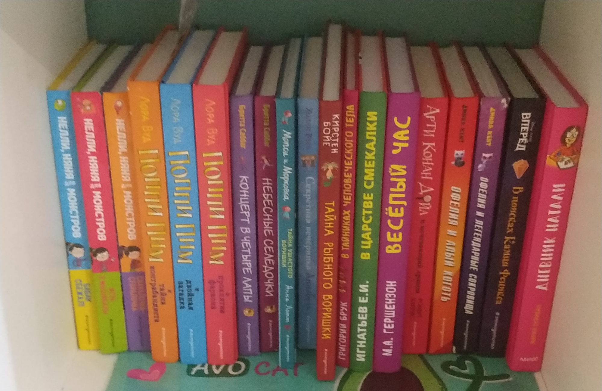 A photograph shows a shelf full of books, arranged vertically. The books are written in Russian. The spines of the books display various titles and author names. Most of the books are brightly colored in shades of pink, red, orange, purple, blue and green. The photograph is a close-up, with only the bookshelf visible. The image's location is not discernible.