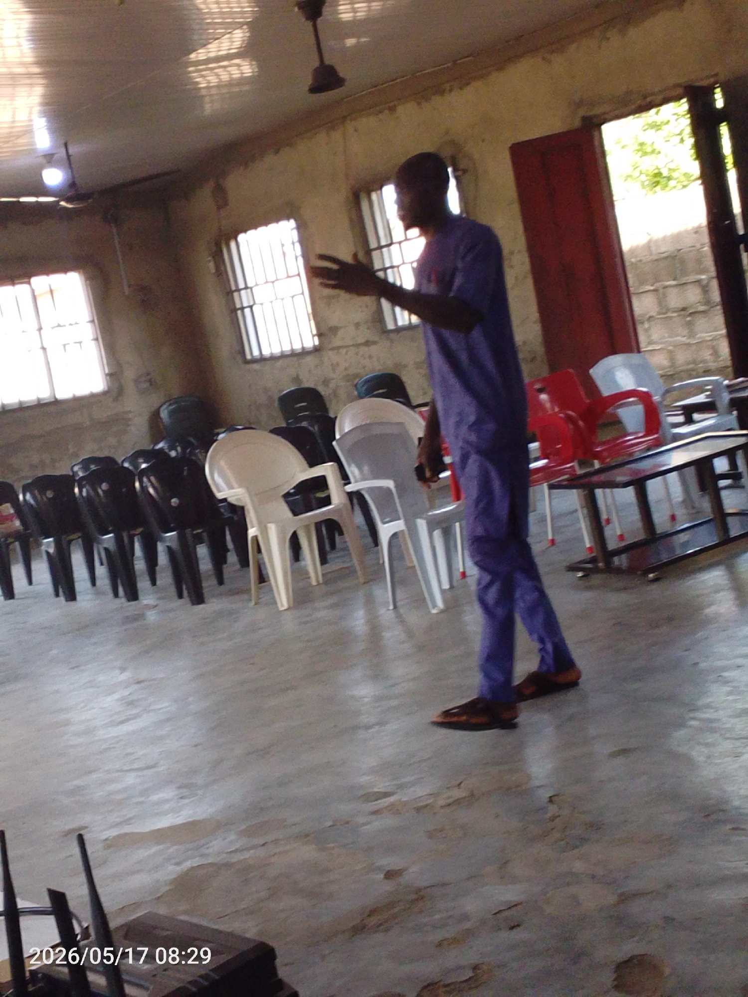 The image shows a room in what appears to be Lokoja, Nigeria, indicated by the surroundings and local appearance of the individuals. In the center of the frame, a man is walking and gesturing. He is wearing a matching top and pants in a shade of blue or purple, and brown leather-look sandals. He appears to be inside a hall or communal space.
Several rows of plastic chairs are arranged in the room, predominantly black and white. There are windows with bars on them allowing light into the room. A table with a metal frame is visible on the right side of the image. There is also a doorway open. The flooring is worn. A ceiling fan is visible in the center of the room. The picture includes a timestamp of 2026/05/17 08:29.