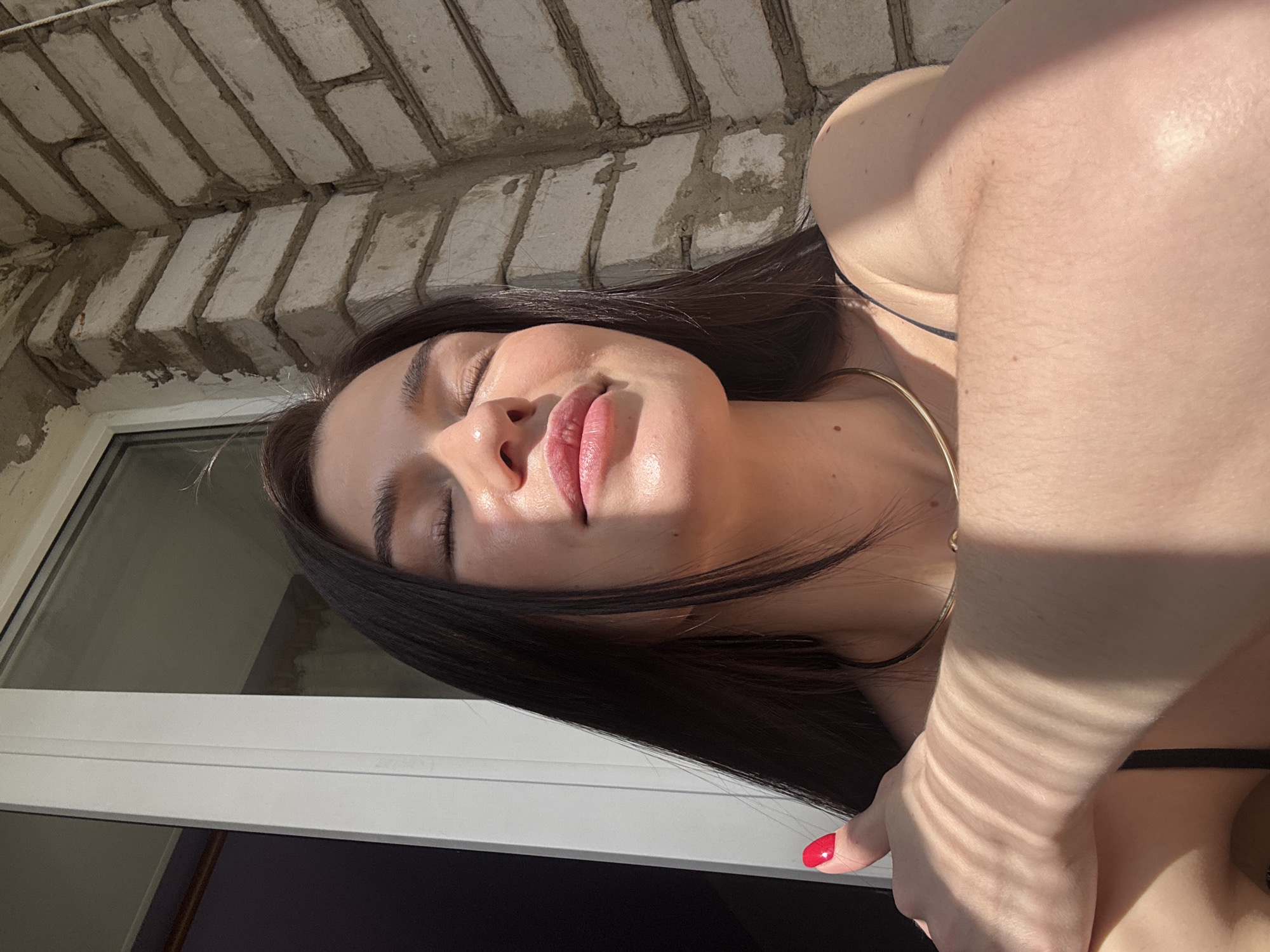 A woman with dark hair lies on her back, eyes closed, face toward the sunlight. She has pink lips and her skin is fair. A gold necklace is visible on her neck. She wears a black strap on her shoulder, and her nails are painted red. Part of a white window frame is visible in the lower left. Above her head, there is a wall made of light-colored bricks. The location is the city of Chaikovsky, Russia.