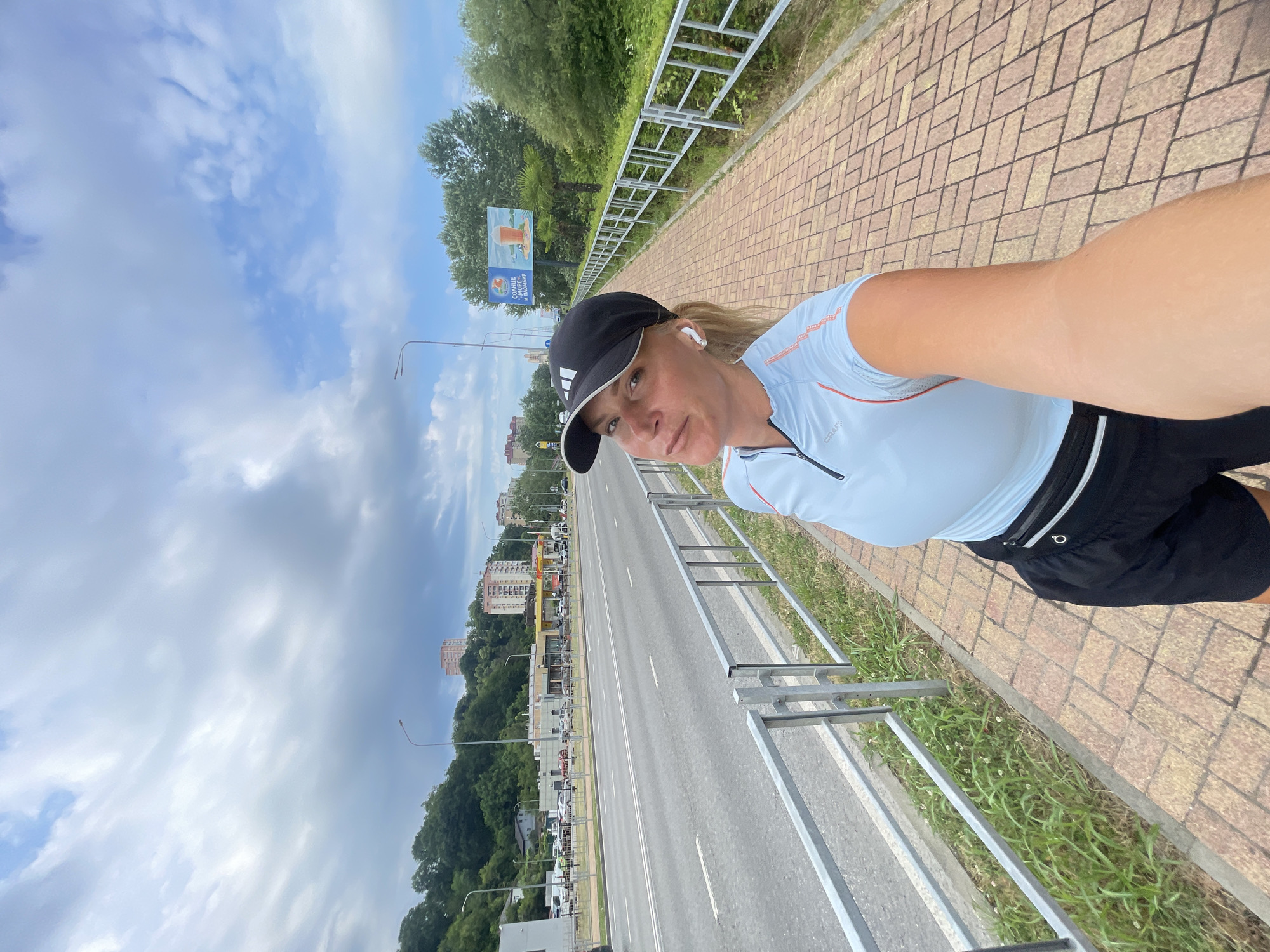 A woman wearing a black baseball cap and a light blue athletic shirt with orange trim takes a selfie while jogging. She has on black shorts and wireless earbuds. She is on a brick sidewalk next to a highway. The highway is lined with a metal guardrail and grassy areas. In the background, there are trees, buildings, and a billboard with an image of a drink. The sky is partly cloudy, suggesting it is daytime. The scene appears to be in an urban or suburban area with a lot of greenery. The woman's expression is neutral. The billboard has text in Russian that says "СОЛНЦЕ, МОРЕ" which translates to "Sun, Sea".