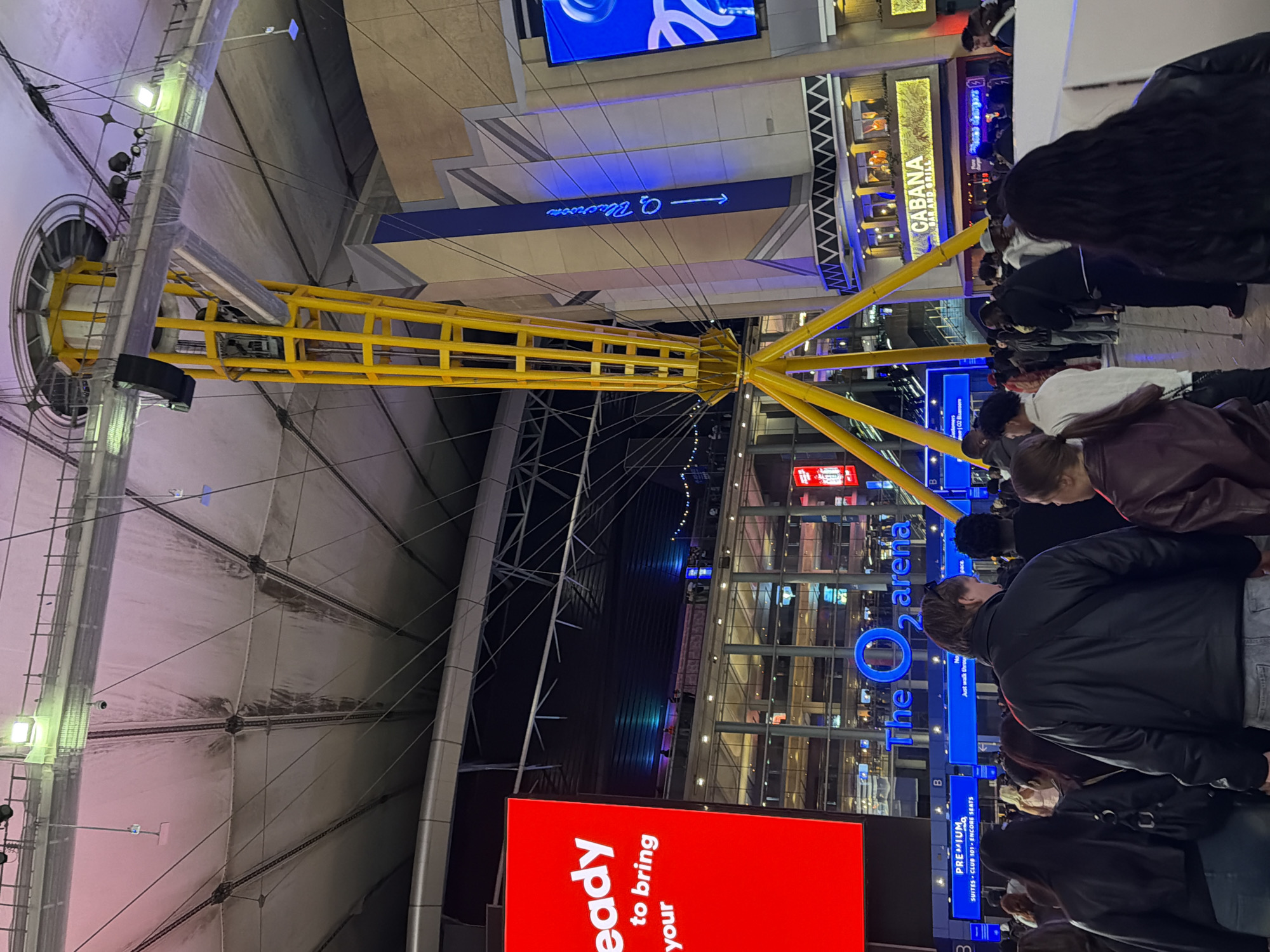 The image captures an interior view of The O2 Arena in London, United Kingdom. A central yellow support structure dominates the frame, its beams radiating outwards. A white domed ceiling looms above, bisected by the yellow beams. The lower portion of the image reveals a crowd of people standing near signs indicating "The O2 Arena" and "Cabana Bar and Grill." A red sign reading "ready to bring your" is partially visible. The lighting is bright, highlighting the details of the architecture and signage.