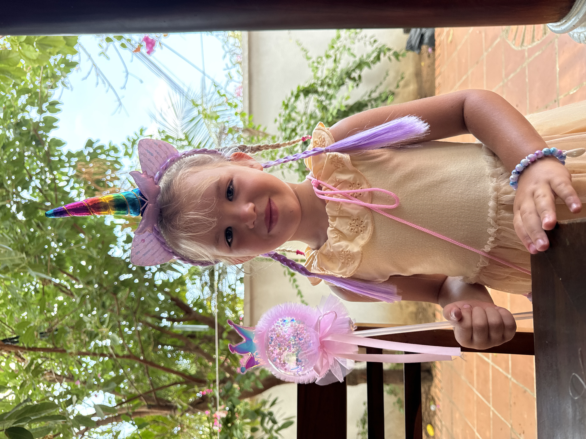 A young girl, likely a toddler or preschooler, is depicted in an outdoor, brightly lit setting. She has light blonde hair, blue eyes, and a gentle smile directed towards the viewer. She is wearing a light peach or beige dress with delicate ruffle detailing and a visible pink string on the bodice.
Her attire is distinctly fantastical; she sports a unicorn headband with shiny purple ears and a vibrant rainbow-colored horn. Complementing this, she has braided hair extensions in a soft purple hue, some with light-colored wraps, that stand out against her blonde hair and rest on her shoulders. On her right wrist, she wears a bracelet composed of marbled blue and pink beads, with a small white charm attached.
In her right hand, she holds a whimsical pink and glittery magic wand. The wand features a clear, sphere-like top filled with sparkling confetti and a small crown motif, adorned with pink ribbons and tulle.
The background suggests a lush, natural environment, likely a garden or patio. Green foliage from trees and climbing plants is visible, creating a vibrant backdrop under a bright, sunny sky. Parts of a terracotta-tiled ground or wall are also discernible, along with a dark wooden beam or frame in the foreground, indicating an outdoor structure. The image appears to be taken in Mũi Né, Vietnam, under clear, daytime conditions. No visible text is present in the image.