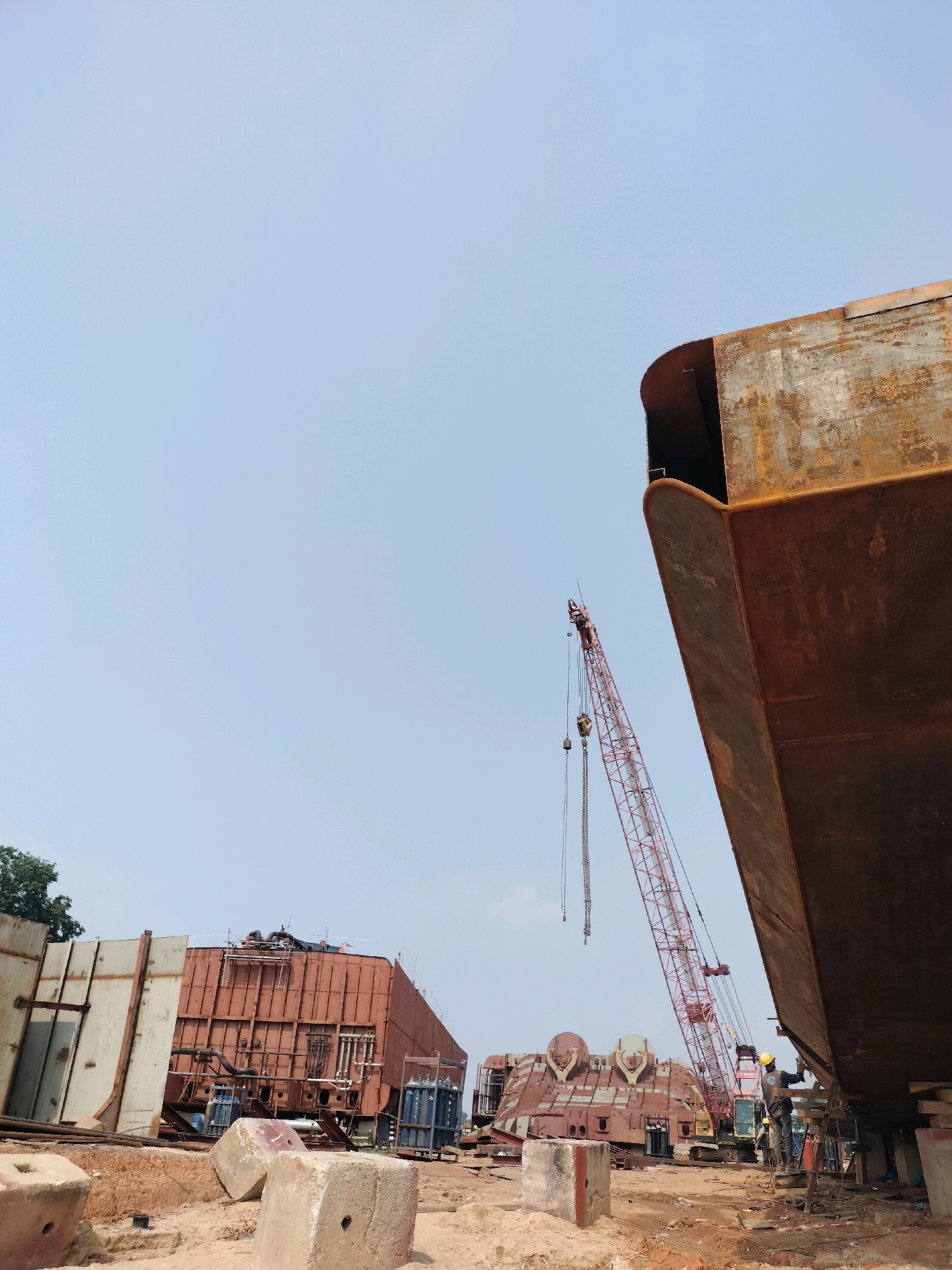 In Batam, Indonesia, a construction site features large, rusty metal ship sections under a clear sky. In the foreground, concrete blocks are scattered on the dirt ground. To the right, the curved hull of a massive ship structure dominates the frame, showing signs of rust and weathering. A red crane with its boom extended is visible, with its hook and chains hanging down. In the middle ground, a large, reddish-brown metal structure, likely another ship component, is situated. Further in the background, more ship sections and construction equipment, including a yellow crane, are present. Several individuals in work attire, some wearing hard hats, are engaged in activities near the ship structures, with one person on a platform appearing to work on the hull in the right foreground. The environment suggests an active shipbuilding or ship repair facility.