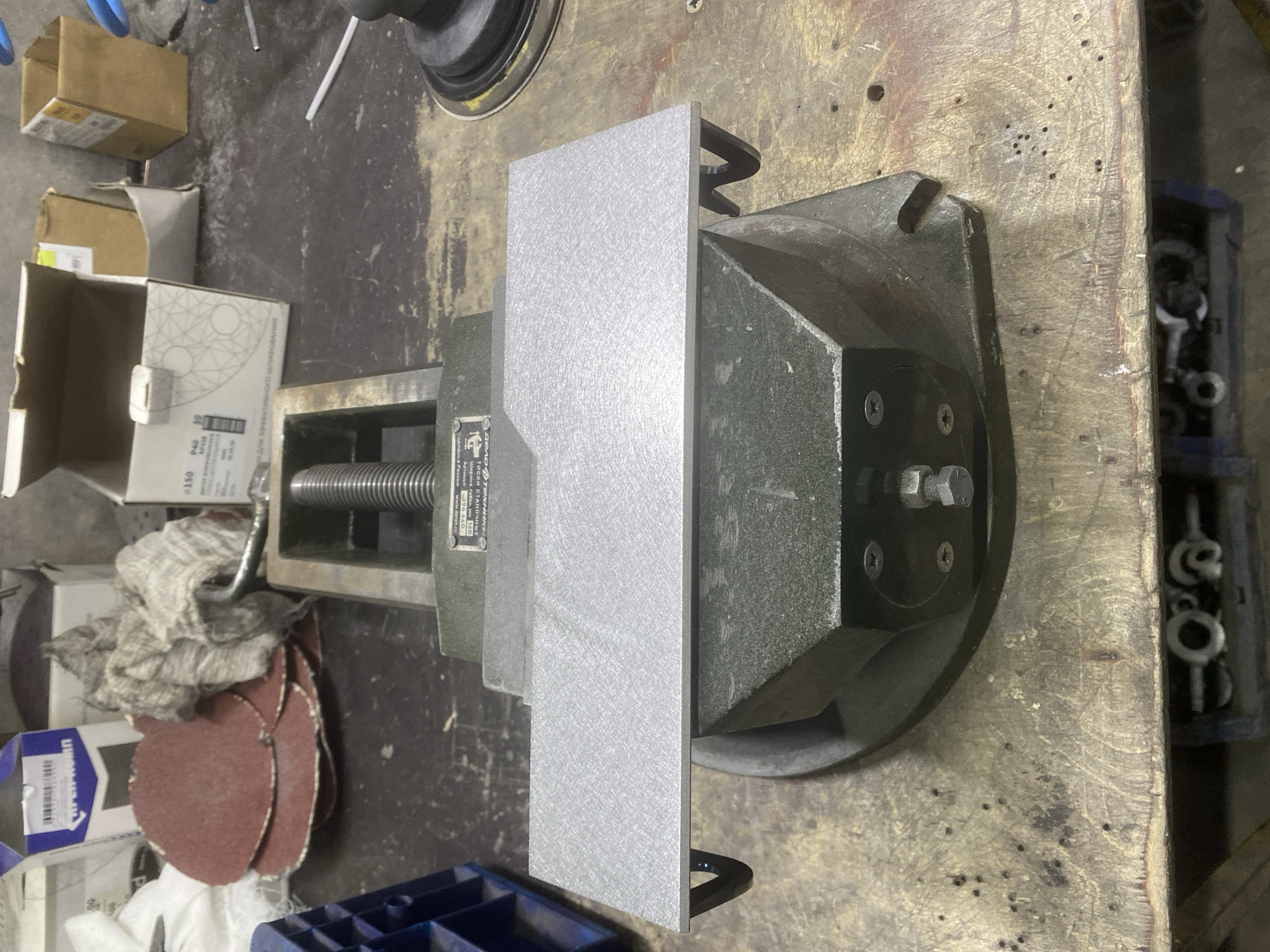 The image depicts a workbench in what appears to be a workshop setting. A metal vise is centrally positioned, holding a flat, rectangular piece of brushed aluminum. The aluminum piece has a slightly angled cut on its right edge. In the background, various items are visible, including cardboard boxes, a bundle of what looks like rags, and a blue plastic container. The workbench surface is worn and appears to be made of wood. The overall impression is one of a functional workspace, possibly used for metalworking or fabrication. There is no indication of the specific location in Berezhki, Russia, nor are there any people or specific actions being performed.