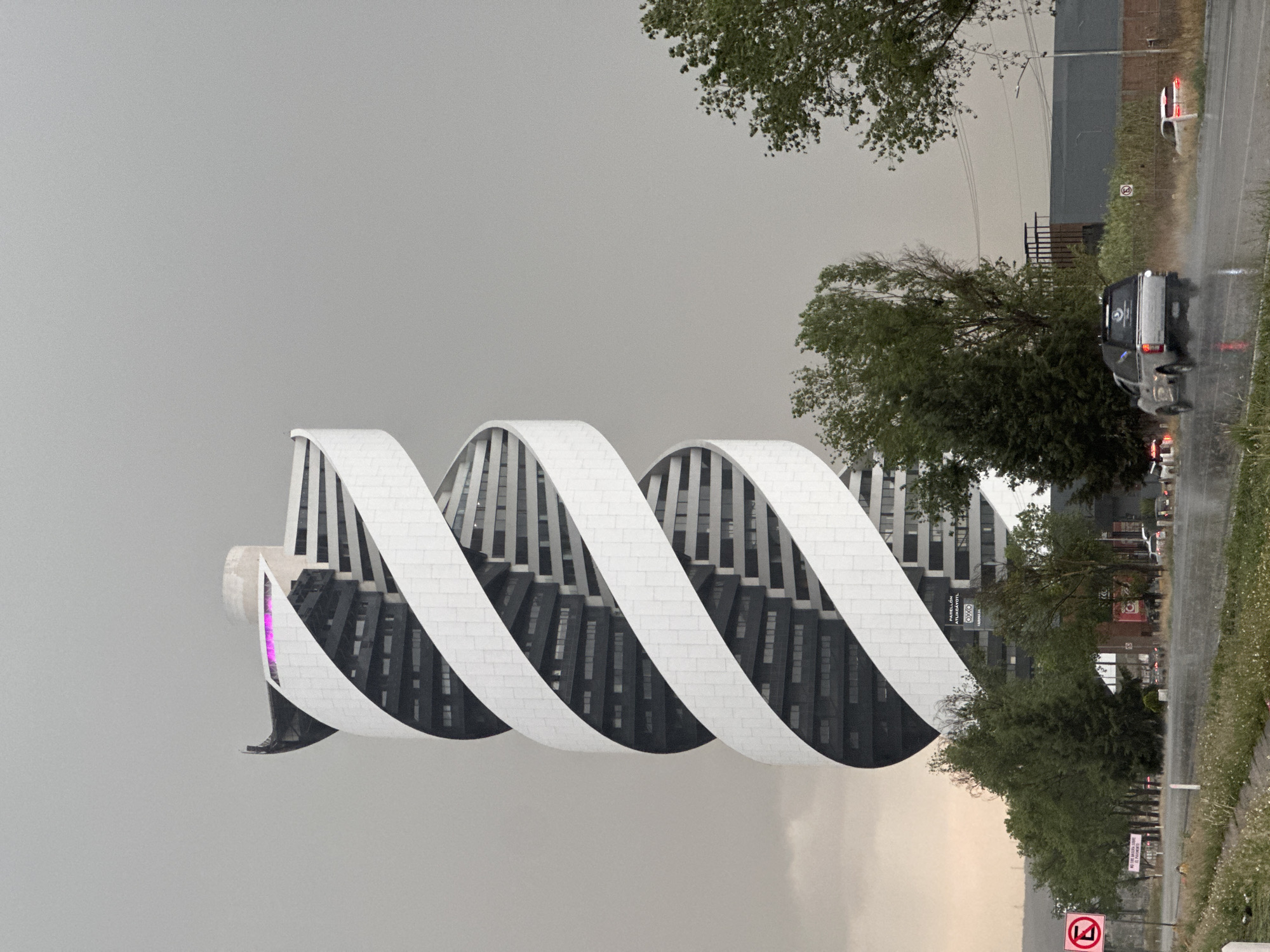 The image depicts the Pabellón Atuxcayotl building in San Bernardino Tlaxcalancingo, Mexico. The building features a unique, spiraling design with white, arching bands wrapping around its dark, multi-storied core. The sky is overcast, suggesting a gloomy day. To the right of the building, there's a road with a few vehicles, including a light-colored pickup truck. Green trees and vegetation line the road. There's also a sign visible near the road. The image appears to have been taken from a vehicle, possibly during light rain.