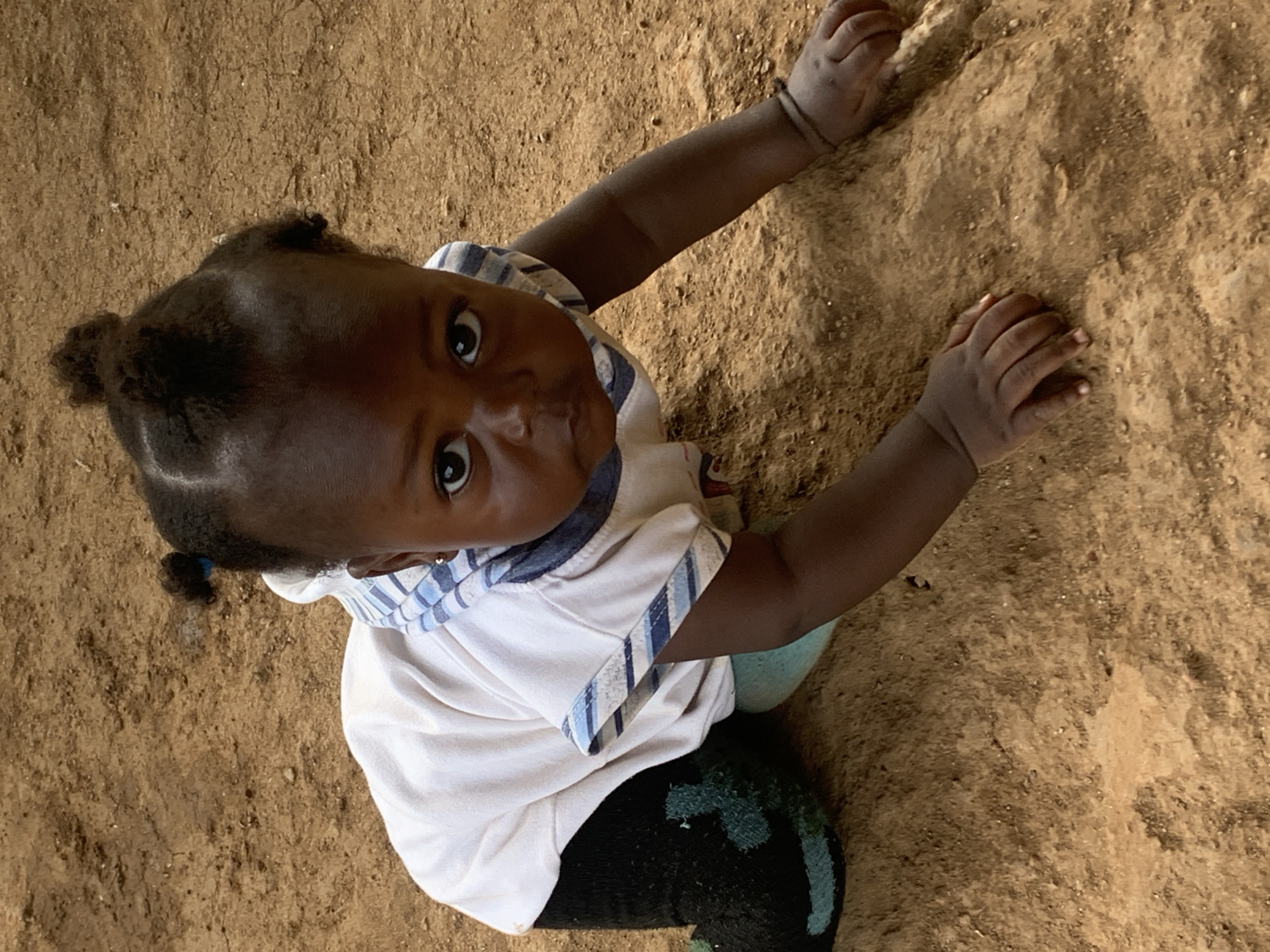 A high-angle, close-up, full-body shot shows a dark-skinned baby crawling on dry, sandy ground, likely in the city of Agban, Nigeria. The baby is wearing a white t-shirt with blue stripes around the collar and sleeve cuffs, and dark pants with greenish accents. The baby has dark curly hair pulled into two small buns and is looking directly at the camera with a serious expression. The baby's hands are on the ground, and the background consists of the brown earth.