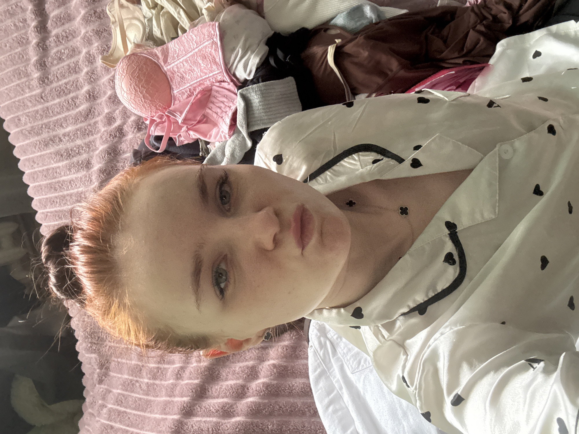 A person is lying on a pink blanket with various items of clothing scattered around. The person has fair skin, red hair tied in a bun, and is wearing a white pajama top with black heart patterns and black piping. A pink corset and other clothing items, including what appears to be a brown garment, are visible in the background. The image does not provide sufficient information to determine a location in Sinyavino, Russia.