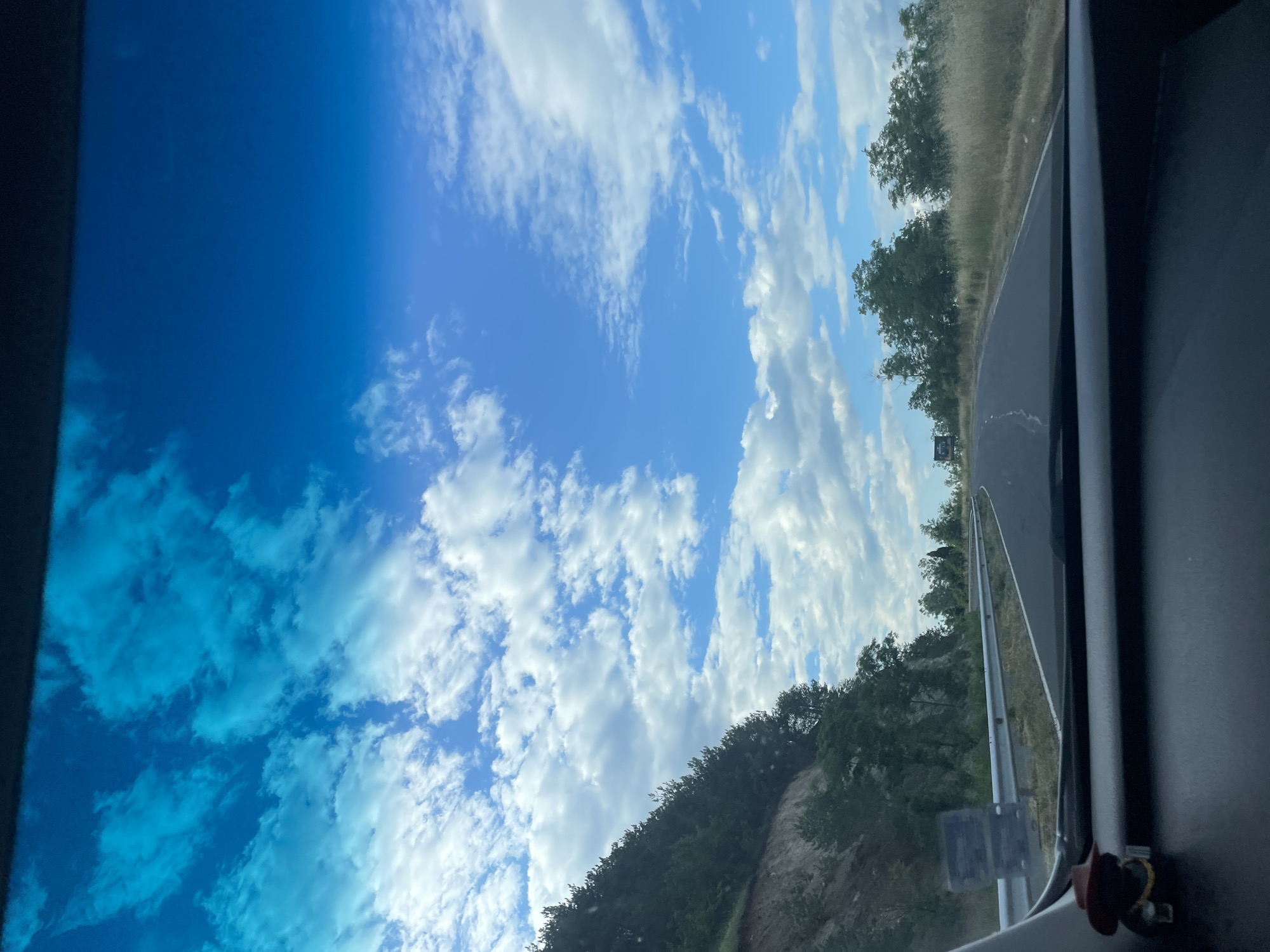 The image depicts a view from inside a vehicle, looking forward through the windshield. The road ahead curves to the right, bordered by a grassy shoulder and a metal guardrail on the left. A steep, wooded embankment rises along the left side of the road, with dense green trees covering its slope. The sky is a vibrant blue, filled with scattered white and light blue clouds. The dashboard of the vehicle is visible in the foreground, with a small, dark figurine wearing a brown hat placed on the left side. The scene appears to be a scenic drive on a highway outside of Balabanovo, Bulgaria, on a clear day.