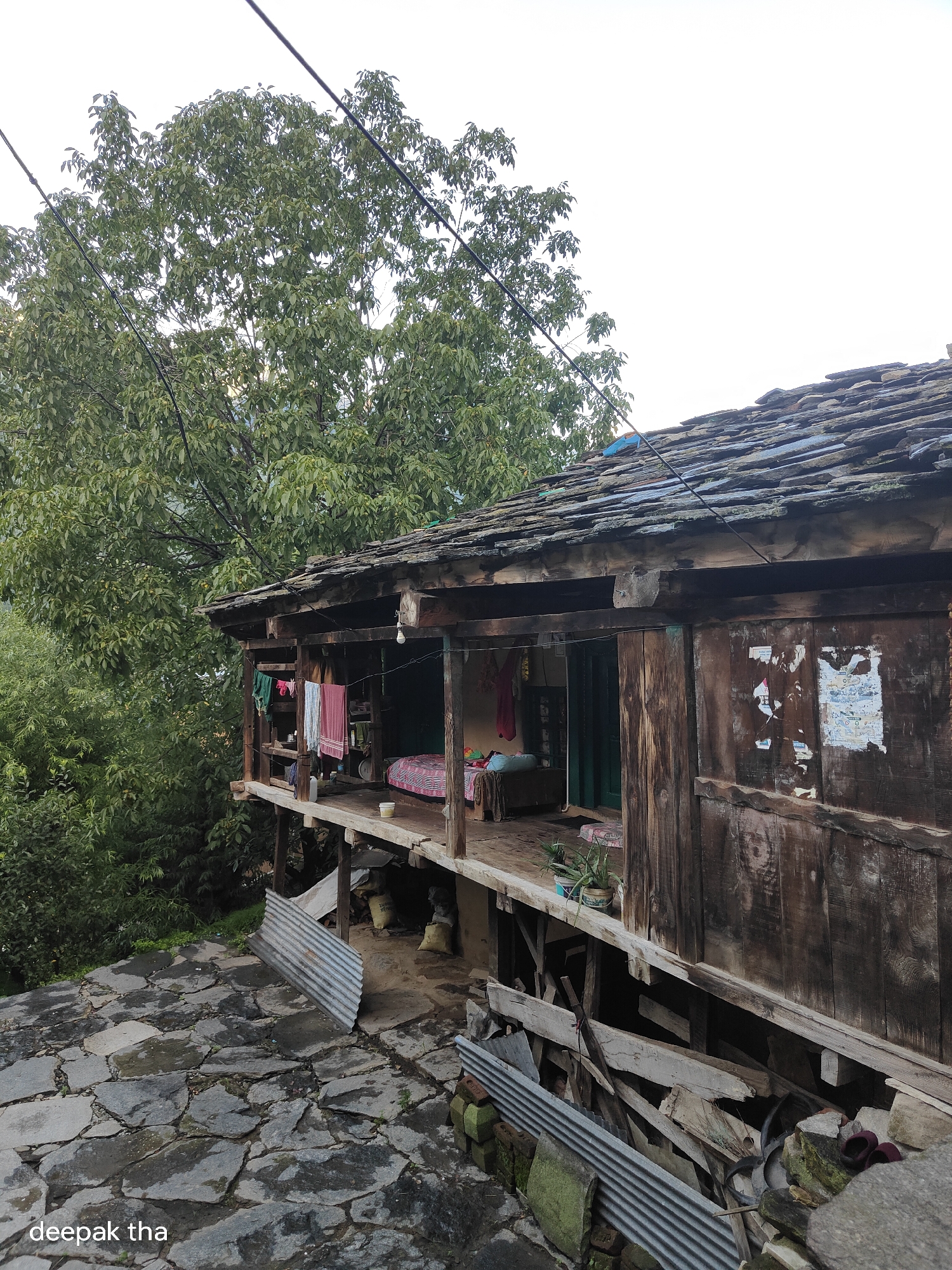 The image depicts a weathered wooden structure with a slate roof, characteristic of traditional architecture in the mountainous region of Vashisht, India. The house is built on stilts, with an open veranda where clothes are hanging on a line, suggesting a domestic setting. The veranda overlooks a stone-paved pathway that appears damp, indicating recent rain or mist, common in the hills. Lush green trees surround the structure, with their dense foliage partially obscuring the sky. Debris, including corrugated metal sheets and wooden planks, is piled beneath the veranda, hinting at ongoing construction or disrepair. The overall impression is one of rustic simplicity and a close connection to nature, typical of rural Indian villages.