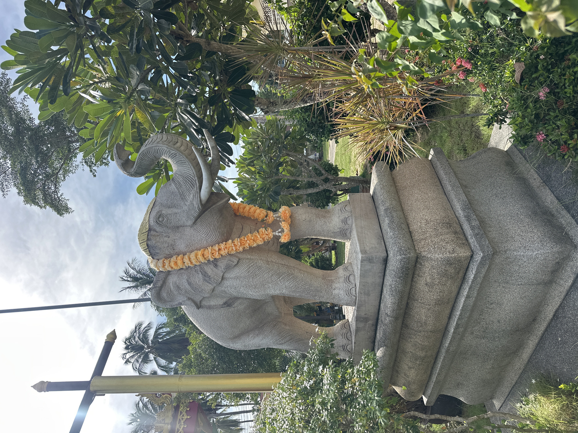 A stone elephant statue stands on a tiered pedestal in a garden on Ko Samui, Thailand. The elephant is decorated with an orange flower garland around its neck. Behind the statue, palm trees and lush green foliage are visible, with a bright sky overhead. A gold-colored pole with a dark horizontal bar is to the left of the elephant. The environment suggests a warm, tropical climate.