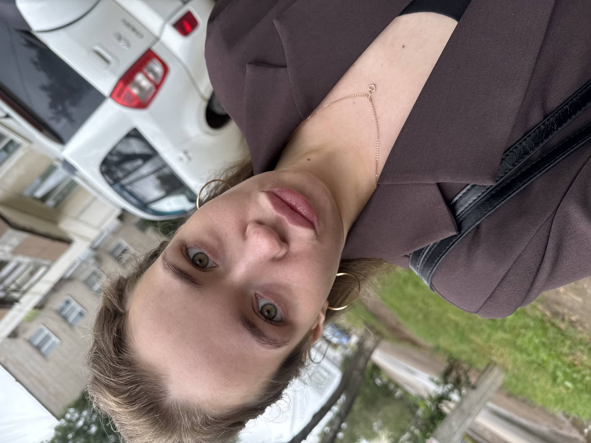 A woman with light brown hair and green eyes is taking a selfie outdoors. She is wearing a brown blazer, a black top, a gold necklace with a small heart pendant, and gold hoop earrings. She is standing in front of a white car and a building. The background suggests an urban setting. The image appears to be taken on a street in Chaikovsky, Russia.