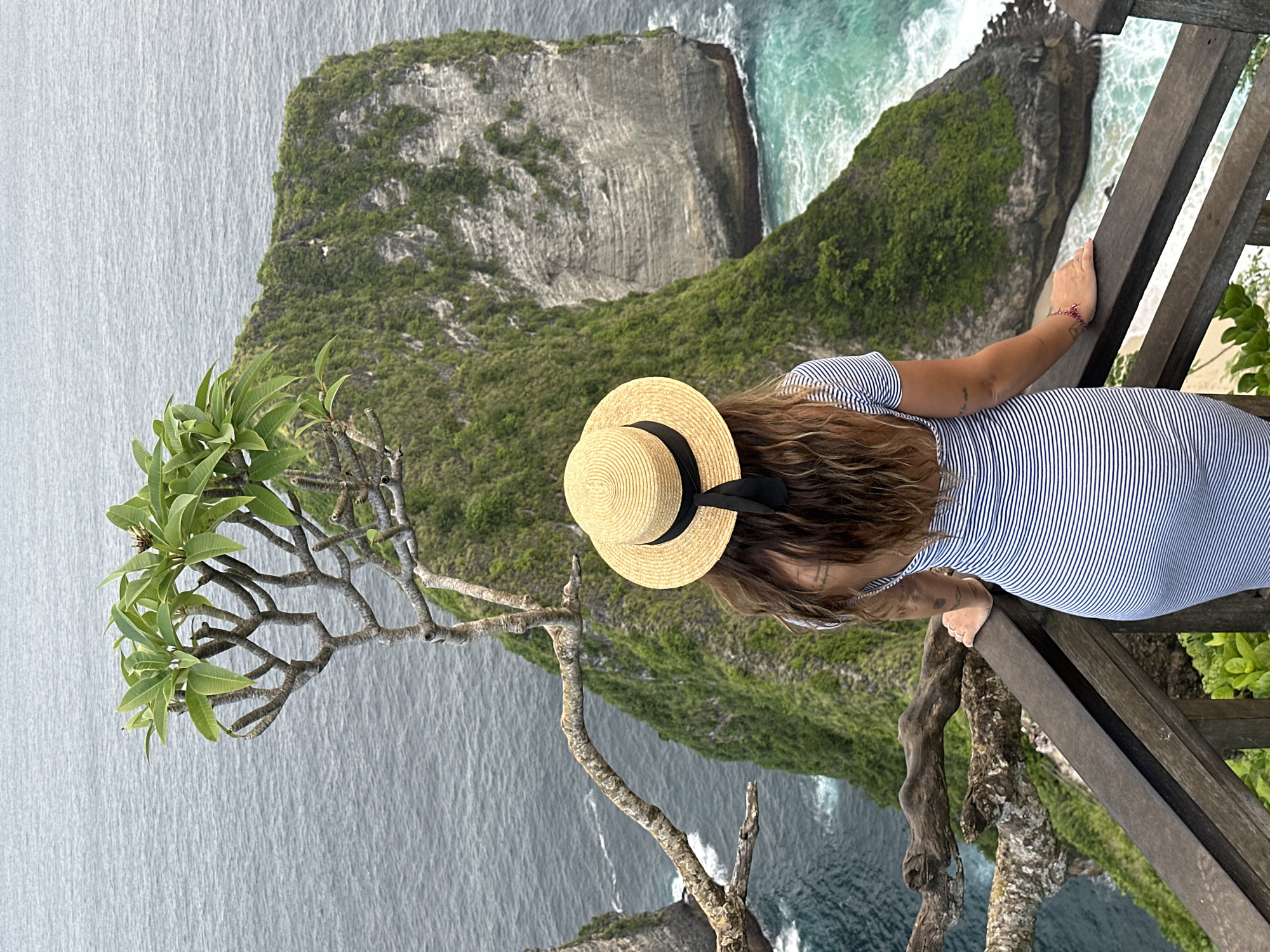 A woman in a striped dress and straw hat stands at a wooden railing, looking out at the coastal view at Kelingking Beach in Nusa Penida, Indonesia. The vista includes a large, green-topped rock formation jutting out into the turquoise sea. The foreground features a tree branch with green leaves, adding depth to the scene. The sky appears overcast, casting a soft light over the landscape.