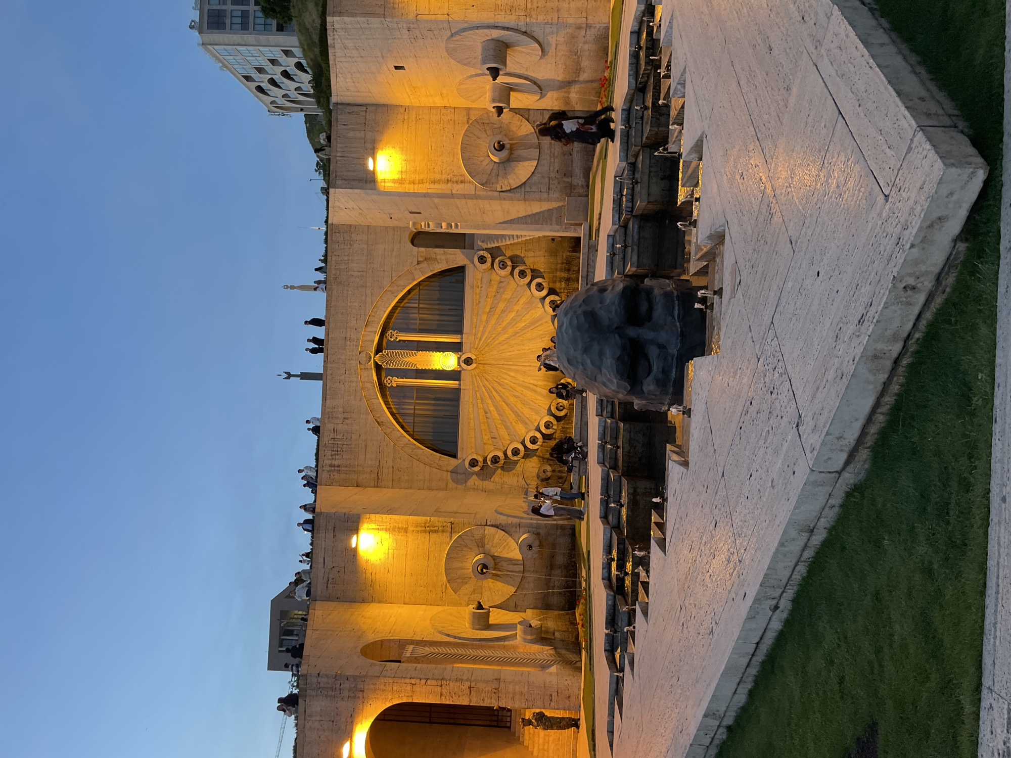 The image shows the Yerevan Cascade in Yerevan, Armenia, at dusk. The camera faces a monument with a large stone head in the foreground. Behind the monument, a grand stairway and water feature cascade down the hillside. The stone structure features circular decorative elements and a large arched opening, with people visible on various levels. The sky is a gradient of blue, indicating early evening. The lighting highlights the stone architecture with warm, golden tones.