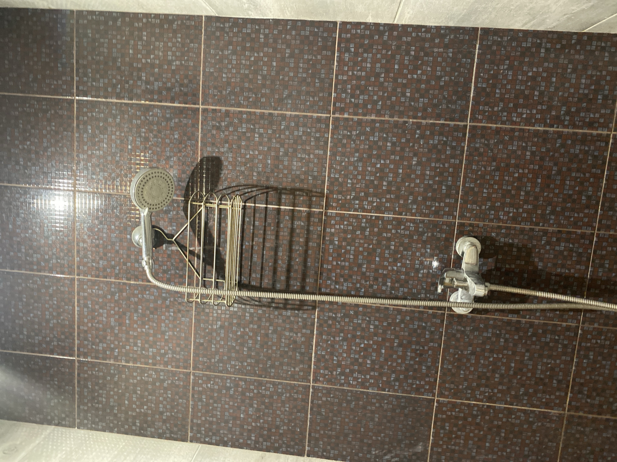 The image shows a close-up view of a shower fixture mounted on a tiled wall. The showerhead, with its characteristic spray holes, is visible at the top, connected to a flexible hose that leads down to a faucet assembly. A metal shower caddy, designed to hold toiletries, is suspended from the shower arm. The wall is covered in small, dark brown and grey square mosaic tiles, creating a textured and patterned surface. To the right of the tiled wall, a lighter colored, larger tile or panel is visible, suggesting a change in material or design in the bathroom. The lighting in the scene creates a shadow of the shower caddy and showerhead on the tiled wall. The image appears to be taken indoors, in what is likely a bathroom. The specific location in Chișinău, Moldova, cannot be determined from the visual information provided in the image.