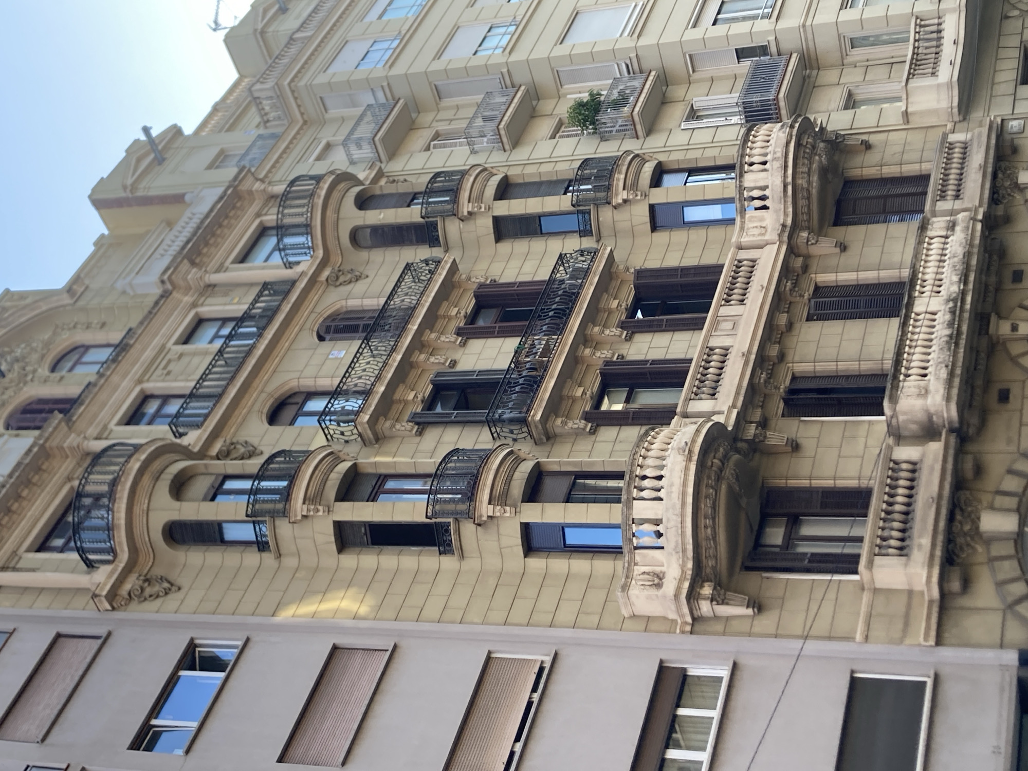 The image displays a close-up, low-angle view of an ornate, light-yellow building in Barcelona, Spain. The building's facade features multiple stories with numerous windows, some of which have decorative black metal balconies. The architecture includes classical elements such as columns, arched window frames, and sculpted details, notably lion heads adorning some of the lower balconies.
The windows themselves vary in design. Many are rectangular and appear to have dark frames and shutters. Some of the upper windows have white blinds. One small balcony on a higher floor contains a single green plant. The building's surface is textured, suggesting it might be clad in tiles or stucco.
Below the ornate building, the top portion of a plainer, grey building is visible. This lower building has simpler, rectangular windows, some of which are open, revealing reflective glass. The overall scene suggests an urban environment with a focus on the architectural details of the older building.
The lighting indicates it is daytime, likely sunny given the reflections in some windows. There are no people or clear activities depicted, making it a static architectural study. The sky is a pale blue, contributing to the bright, daytime atmosphere. No text is visible in the image.