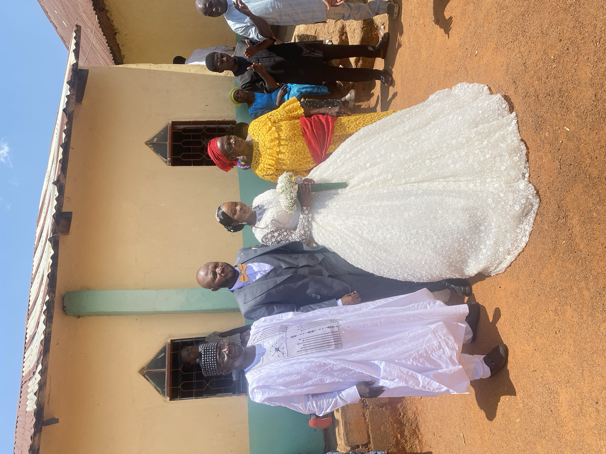 The image captures an outdoor gathering, likely a wedding celebration, in Vom, Nigeria, under bright daylight. A group of people are posing in front of a simple, single-story building featuring light yellow walls, a horizontal light-green stripe, and a corrugated metal roof. The ground is reddish-brown earth.
In the foreground, a bride is prominent in a white, floor-length wedding gown with a textured, patterned overlay, holding a white floral bouquet with green leaves. Her hair is styled, and she wears subtle jewelry. Standing next to her is an older woman in a vibrant yellow dress with a red sash around her waist and a matching red headscarf.
To the right of the bride (viewer's left in the corrected orientation), the groom is seated, or his lower half is obscured, wearing a dark suit, a white shirt, and a yellow bowtie. Next to him, an older man stands, dressed in a long, flowing white traditional tunic (possibly an agbada) with intricate embroidery on the chest and a patterned cap.
Several other individuals are visible in the background: a young woman in a blue dress and a person in a black shirt stand behind the woman in yellow. Further back, near the building, two men are visible, one in a light blue shirt and another in a black shirt. The building has windows fitted with dark security bars.
The bright sunshine creates distinct shadows on the ground, suggesting it is midday or early afternoon. The sky is clear blue. The overall atmosphere appears celebratory. There is no visible text in the image.