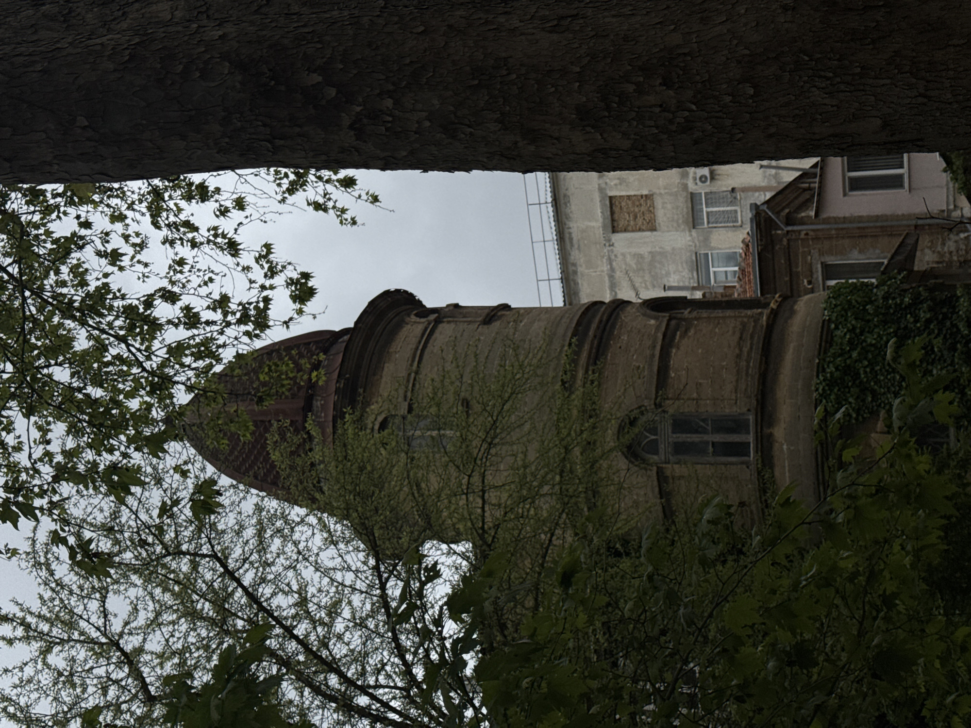 A low-angle shot depicts an old tower with a dome roof, likely a part of a building, seen through the branches of a tree and beneath a large tree trunk that forms the top border of the image. The tower, constructed from light brown stone, has multiple levels or segments and arched windows. Adjacent to the tower is a taller, more modern building with lighter colored walls and visible windows. The sky is overcast. Based on visual features, the location could be Yevpatoriya, Ukraine.