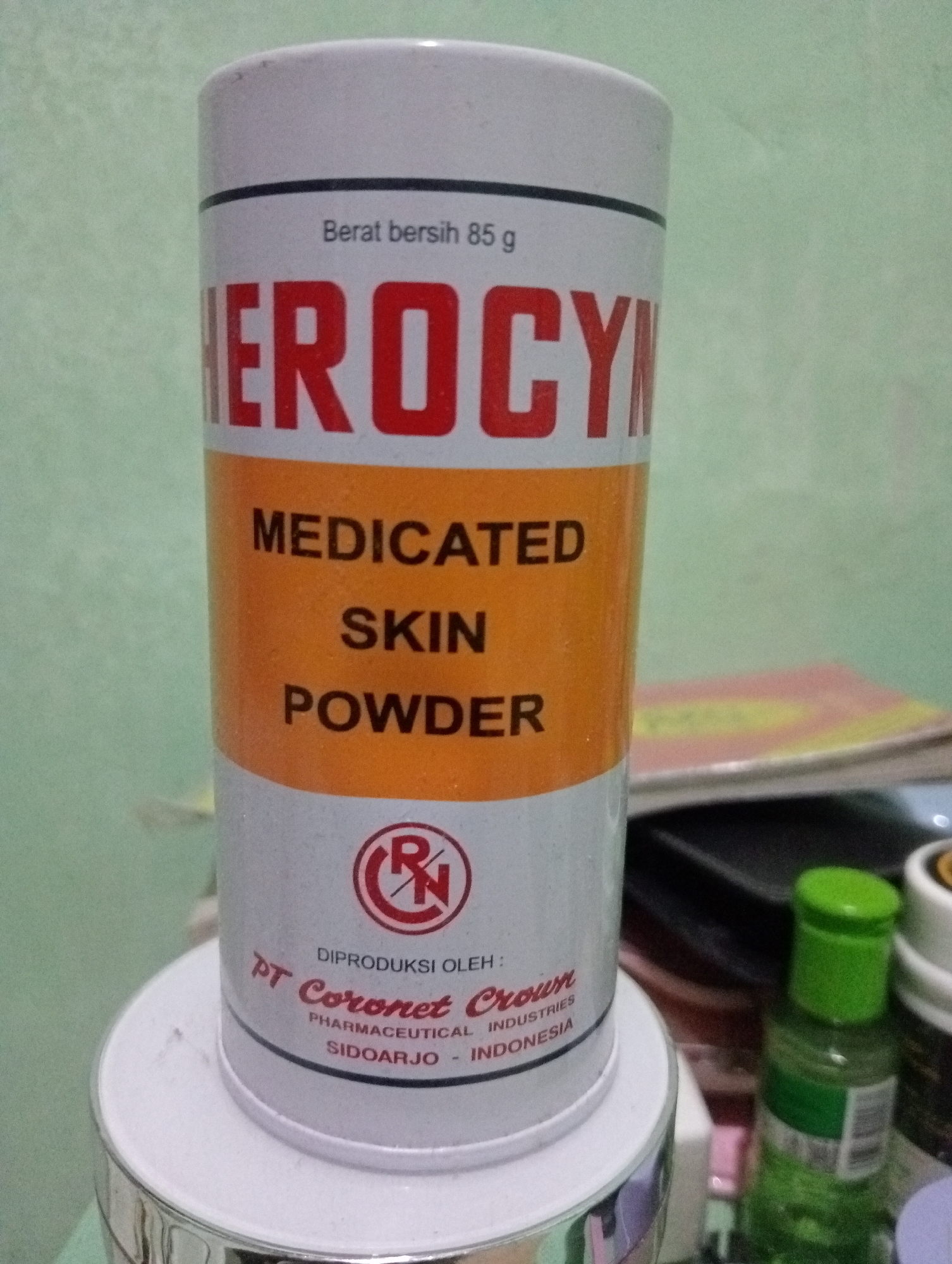 A white and orange cylindrical container of "HEROCYN" medicated skin powder is centrally positioned in the foreground. The container has red text for the brand name and black text for other product details. Below the brand name, the words "MEDICATED SKIN POWDER" are visible in black text on an orange background. Towards the bottom of the container, there is a red and white logo, followed by text indicating it is "DIPRODUKSI OLEH: PT Coronet Crown PHARMACEUTICAL INDUSTRIES SIDOARJO - INDONESIA". The container sits on a white circular base, which is resting on a larger silver object. In the background, to the right of the container, is a green capped bottle and other out-of-focus objects, including what appears to be papers or books. The background wall is a light, muted green. The image was captured indoors. The text "SIDOARJO - INDONESIA" suggests a location in Sidoarjo, East Java, Indonesia, which is not within Kota Negara Timur.