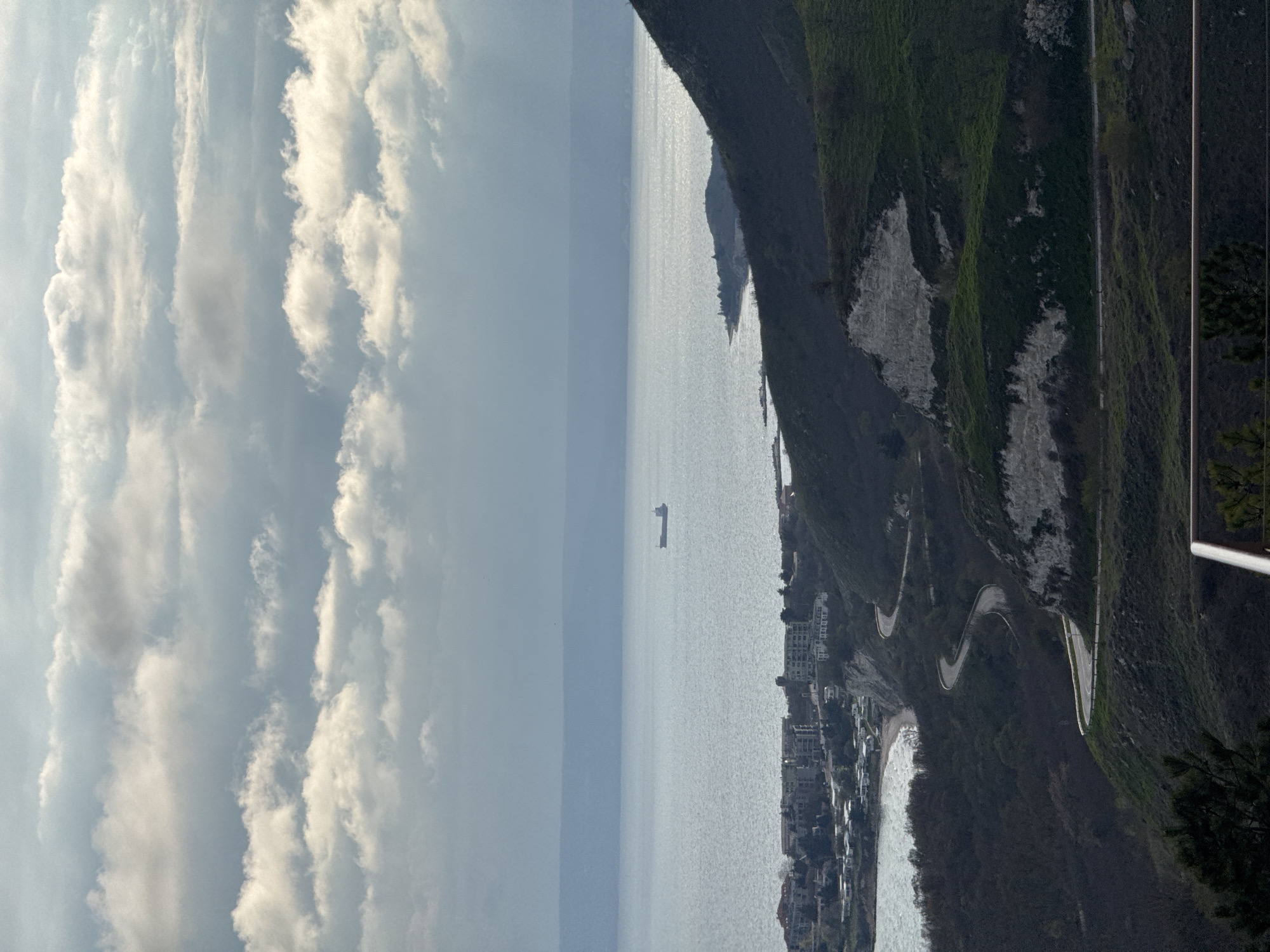 The image shows a coastal scene. On the right is a green, rocky hill with a winding road running along its slope. At the base of the hill, near the water, lies a town with buildings clustered together. In the distance, a cargo ship is visible on the water. The left side of the image is dominated by a cloudy sky.
This scene is not Topola, Bulgaria. Topola is not a coastal city, and the terrain does not match that of the surrounding region.