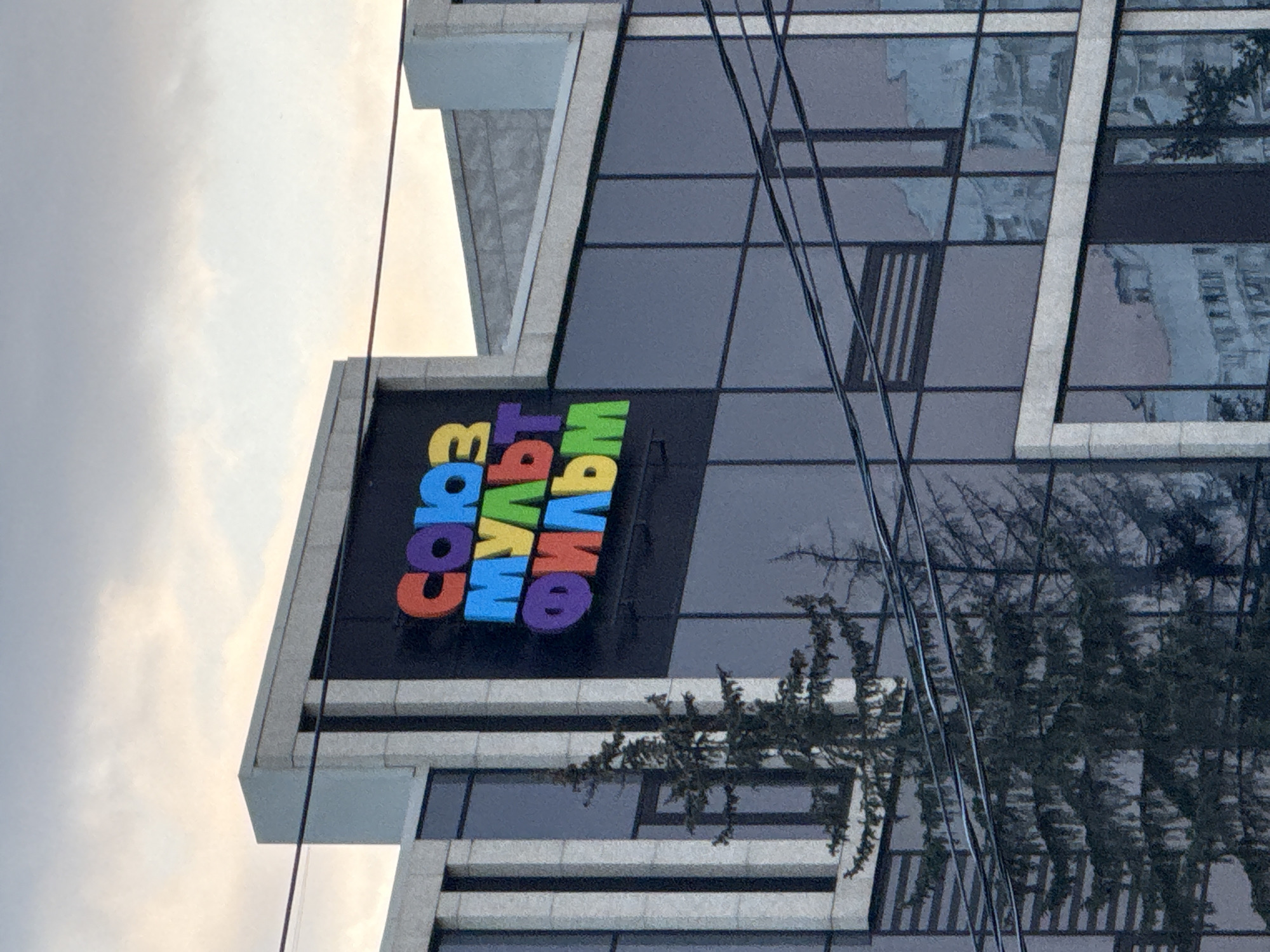 A low-angle shot captures a section of a modern building, identified by the sign "Союзмультфильм" (Soyuzmultfilm) in colorful, block letters. The building, situated in Khimki, Russia, features a gray façade with dark, mirrored windows. The reflections in the glass show the silhouettes of trees. Some overhead wires partially obstruct the view of the building. The sky above is overcast with gray clouds.