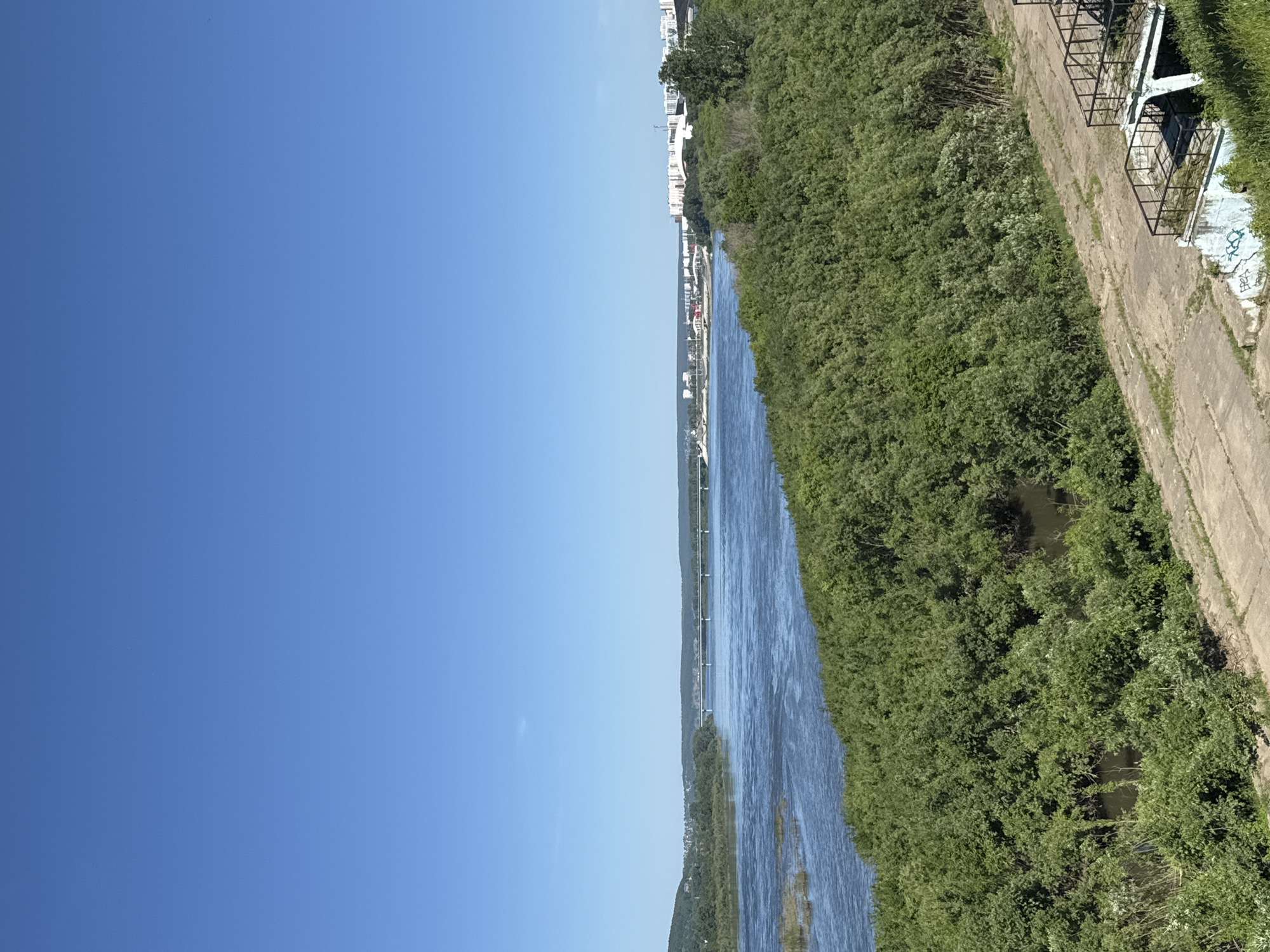 This media file depicts a wide, blue river flowing beside a dense, green forest. The sky is clear and bright blue, indicating a sunny day. The river reflects the sky and is slightly rippled, suggesting a gentle current. On the right side of the riverbank, a paved path runs alongside the trees, with some concrete structures and what appear to be remnants of a fence or barrier. The vegetation is lush and full, with tall trees and undergrowth. In the distance, on the opposite bank, there are glimpses of buildings, suggesting a town or city located there. The perspective is from a higher vantage point, looking down and across the river. The overall scene conveys a sense of nature's tranquility with a hint of urban proximity. There is no visible text in the image.