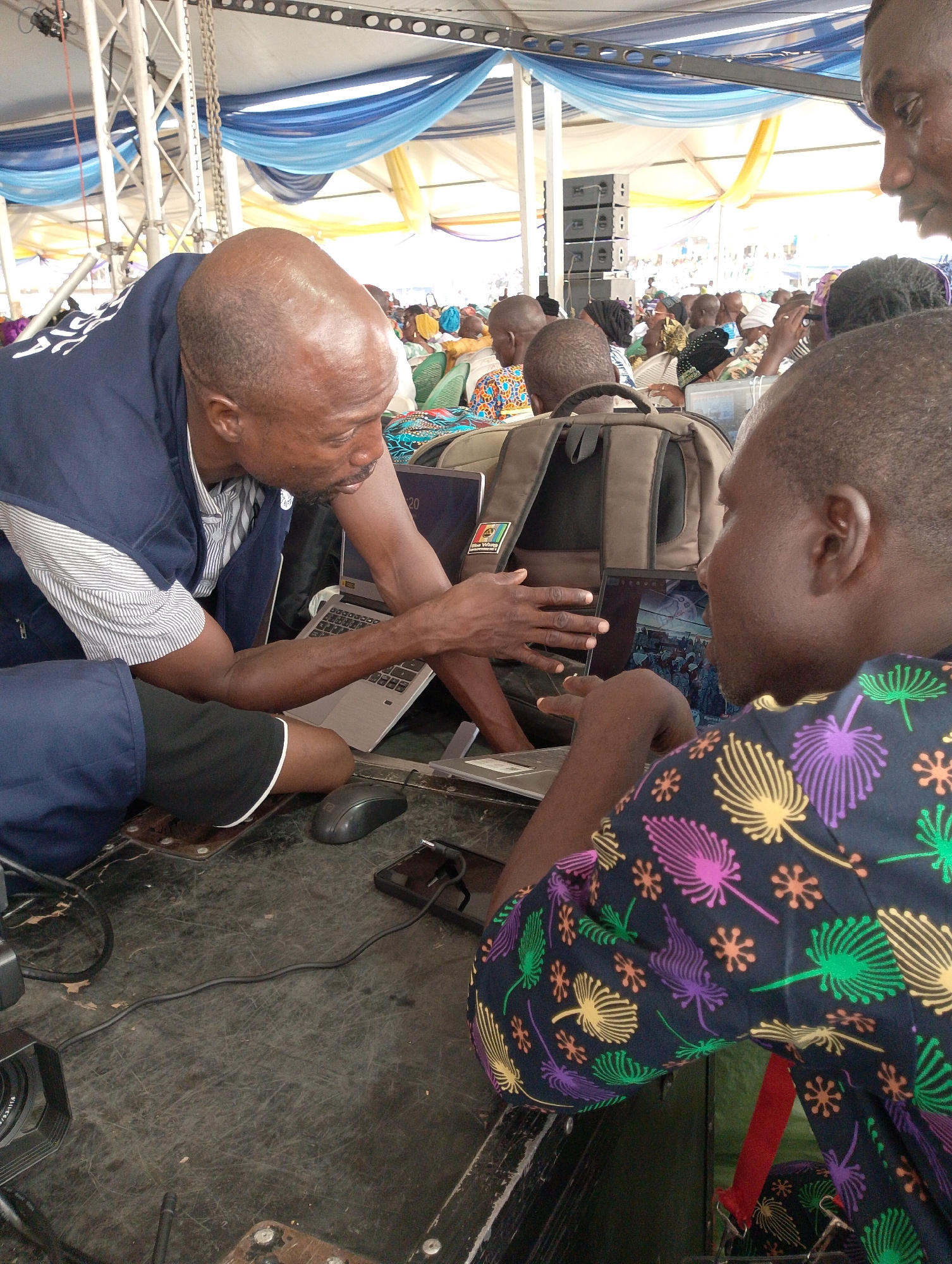 The image shows two individuals interacting over a laptop in an outdoor setting, presumably in Dushepe, Nigeria, due to the patch on the backpack. One man, who is bald and wearing a striped shirt and blue vest, is pointing at the laptop screen. The second man, wearing a dark shirt with a colorful floral pattern, is also looking at the screen. A mouse, a cell phone connected to a charging cable, and a camera lens are visible on the surface in front of them. In the background, a crowd of people is seated under a structure adorned with blue and yellow fabric. The setting appears to be an event or gathering.
