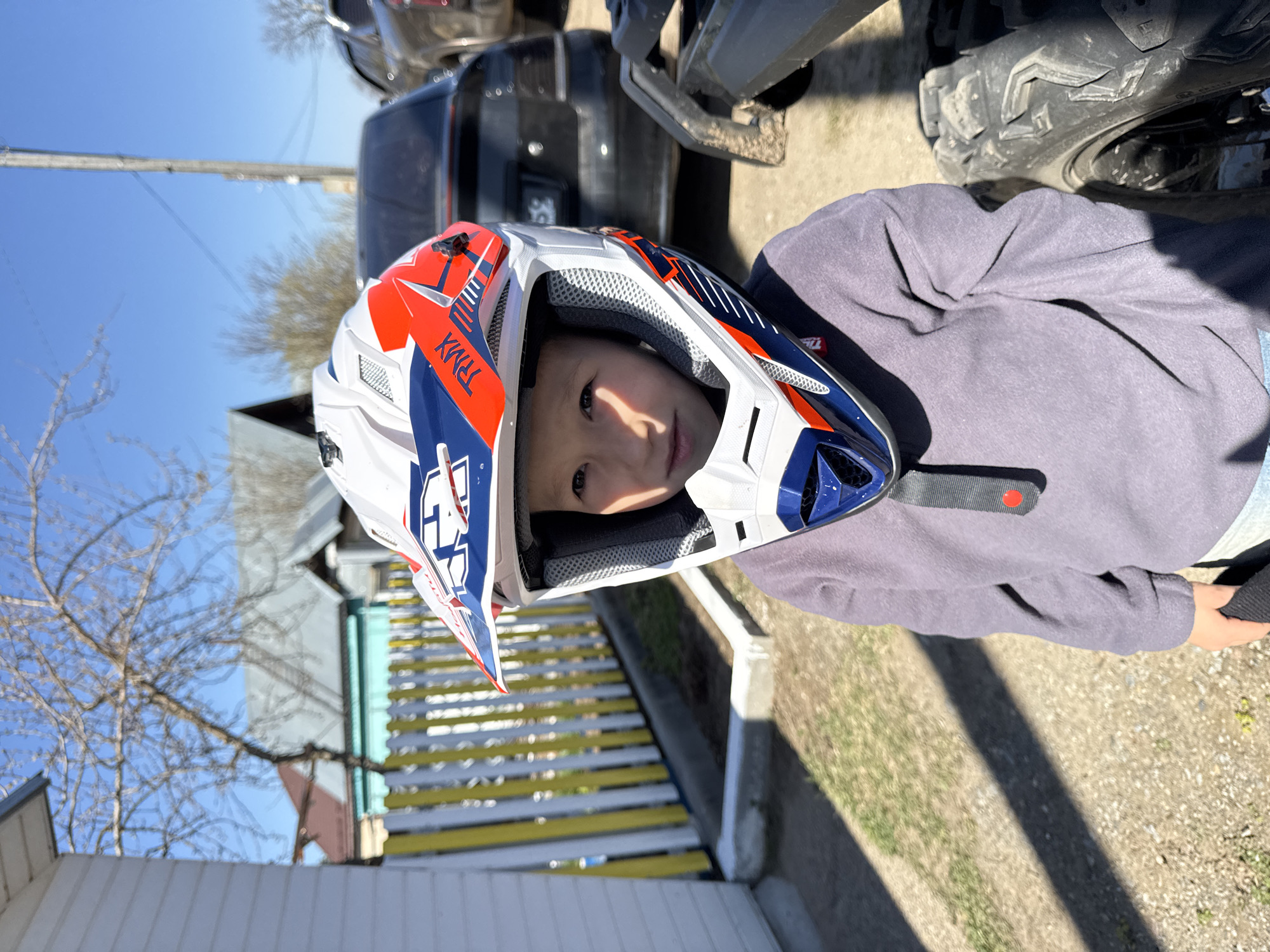 A child wearing a white, red, and blue motocross helmet is the primary subject of the image. The child appears to be standing outdoors near an ATV, a dark SUV, a yellow and white fence, and a house. Bare trees are visible in the background. The photo may have been taken on a residential street in Chaikovsky, Russia.