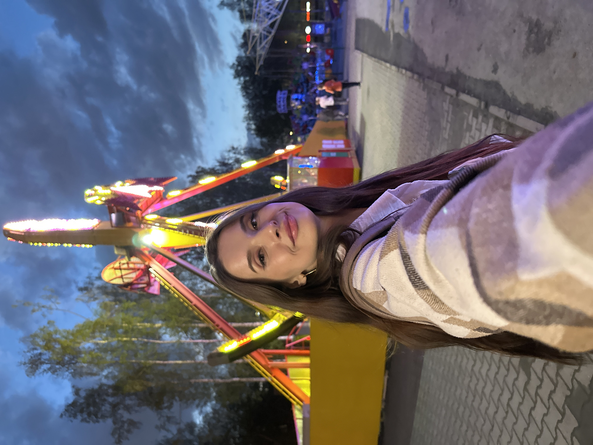 A young woman takes a selfie in a fairground in the city of Yekaterinburg, Russia. She is facing the camera, with a fairground ride behind her, brightly illuminated in the evening light. The sky above is a deep blue, with dark clouds visible. Other attractions and people are in the background, and the ground is paved.